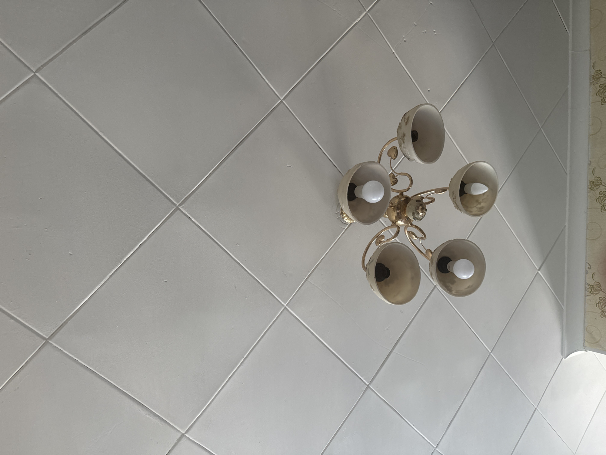 The image shows a close-up, low-angle shot of a white, tiled ceiling with a gold chandelier hanging in the center. The ceiling is divided into diamond-shaped tiles by recessed lines. The chandelier has five arms, each with a beige-colored lampshade and a visible light bulb. The top of the image is dominated by the ceiling, with the chandelier occupying the lower half. The lighting suggests an indoor environment, likely a room in a residential building. There are no people visible in the image. The location in Aqköl, Kazakhstan is not discernible from the visual information in the photograph.