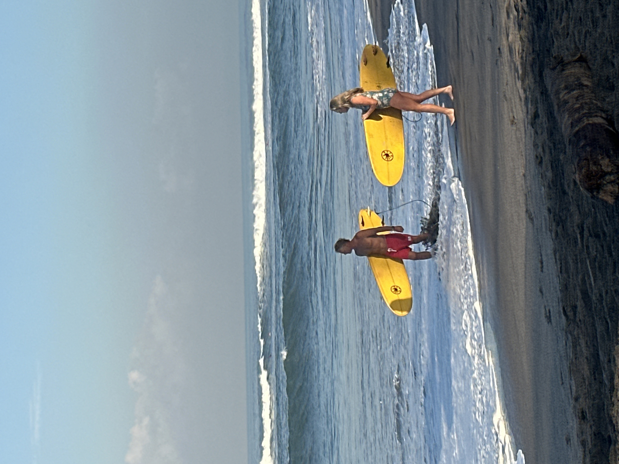 Two surfers, a male and a female, walk along the shoreline of a beach in North Kuta, Indonesia, carrying yellow surfboards. The male surfer, with a muscular build, wears red swim trunks and stands in shallow water, while the female surfer, in a patterned swimsuit, walks on the wet sand. The ocean behind them features gentle waves breaking on the shore, with a clear blue sky overhead. The beach is dark sand, with a piece of driftwood visible in the foreground.