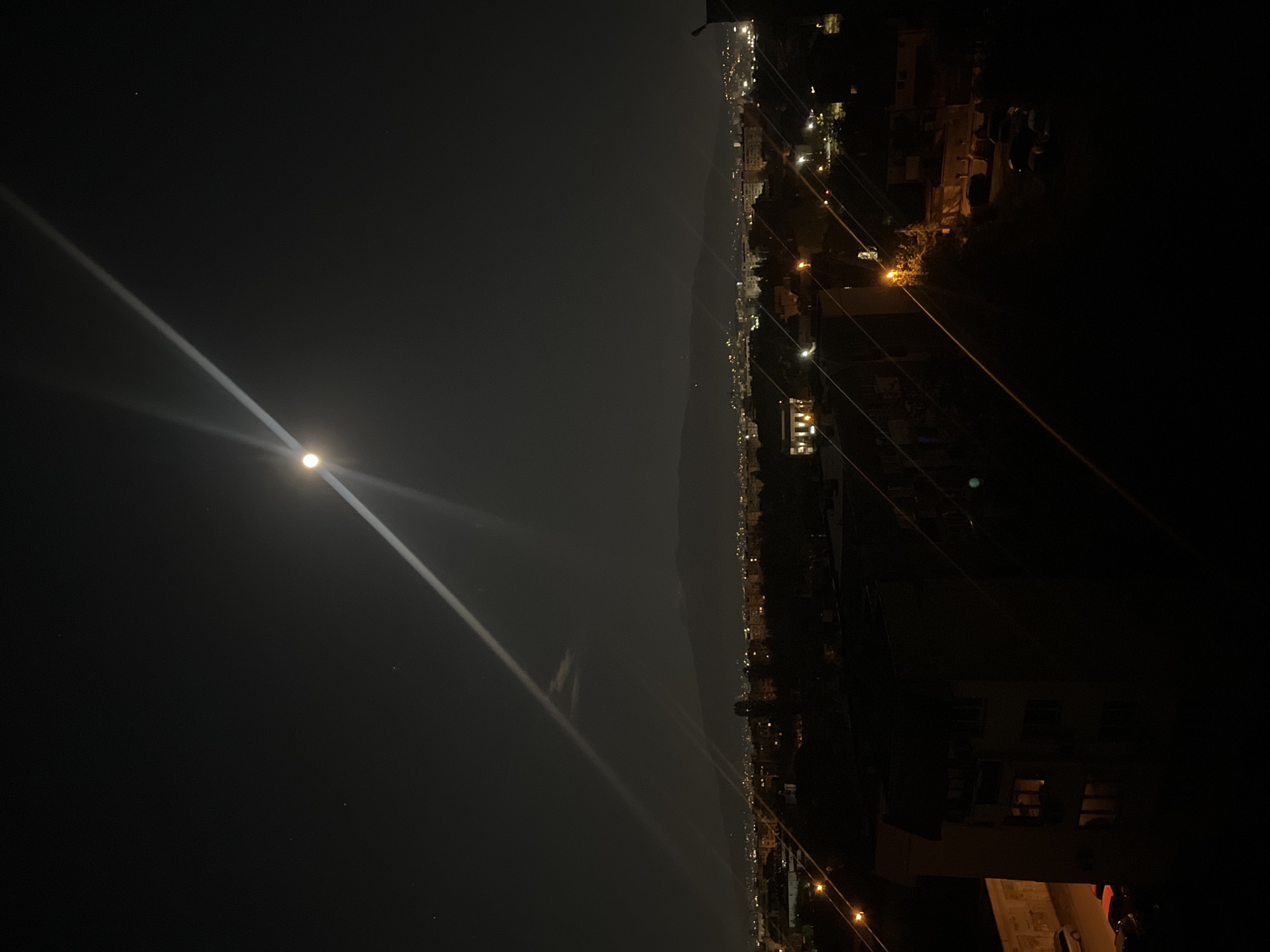 A dark night cityscape in Sofia, Bulgaria, oriented horizontally. The moon is visible on the left, emitting a bright light ray across the scene. In the center, a distant mountain range is silhouetted. Below the mountains, city lights illuminate the terrain. The right side of the image is dominated by dark shadows, with glimpses of buildings, streetlights, and power lines. There are no people directly visible in the image.
