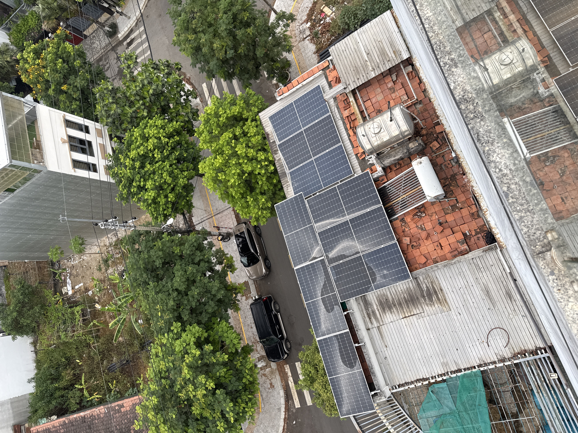 The image, taken from an elevated perspective, showcases a street scene in Đà Nẵng, Vietnam. The street is lined with trees and buildings. Two cars, one black and one silver, are parked along the street. The rooftops of buildings in the foreground have solar panels installed. Other rooftop features include water tanks and a mixture of corrugated metal and tile roofing materials. The area around the buildings includes some foliage and debris. A white building is visible across the street. Crosswalks mark pedestrian pathways. Power lines stretch across the street.
