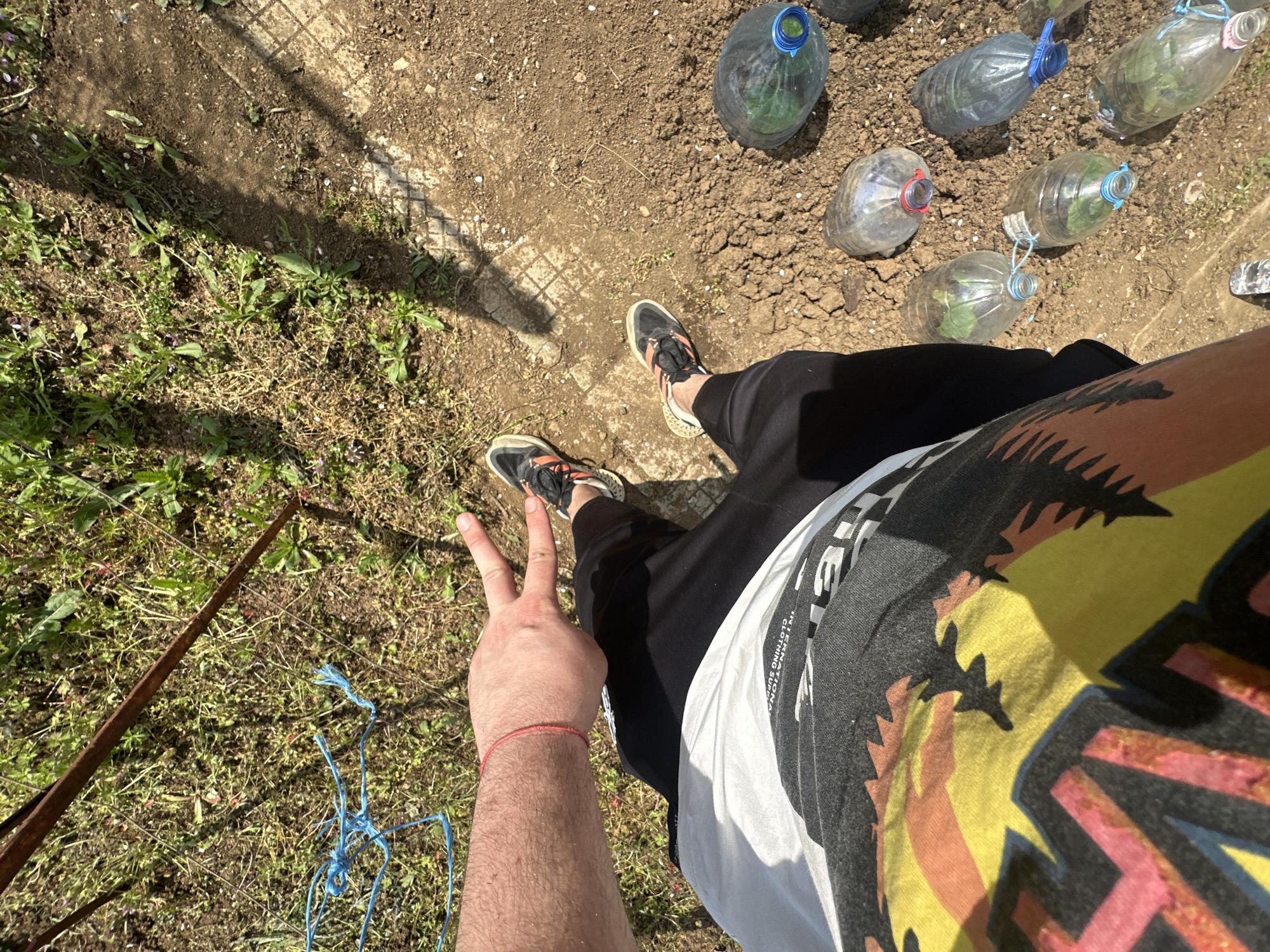 The image shows a person standing in an outdoor area, likely a garden or yard. The location is in the city of Montana, Bulgaria. The person is wearing black sweatpants and a t-shirt with a colorful graphic design over a white undershirt. They are also wearing athletic shoes. The person is holding up two fingers in a "peace" or "victory" sign. A red string bracelet is visible on their left wrist. The ground around the person is dirt and grass. There are several plastic bottles scattered on the ground, some of which appear to be used for growing plants. A rusty metal rod lies on the grass to the left of the person. Blue string is tangled near the metal rod.