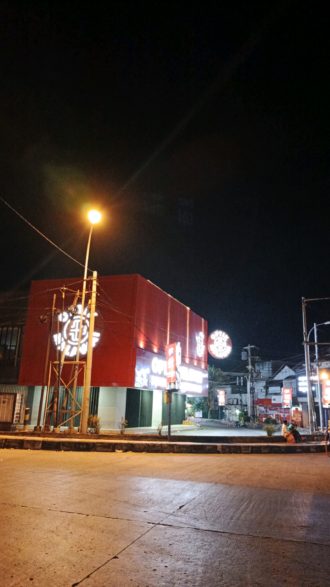 The image depicts a street scene at night in Surakarta, Indonesia. A prominent red building with illuminated signage, "OPTIC SWAYASA," is the central subject. A street lamp with a bright orange light stands in front of the building, casting light on the surrounding area. Several smaller illuminated signs are visible on the building and in the background. Two individuals are seated on a curb in the middle ground, to the right of the red building. The street in the foreground is paved and cracked, with shadows cast by the artificial lighting. The overall atmosphere is one of a commercial area at night, illuminated by artificial light against a dark sky.