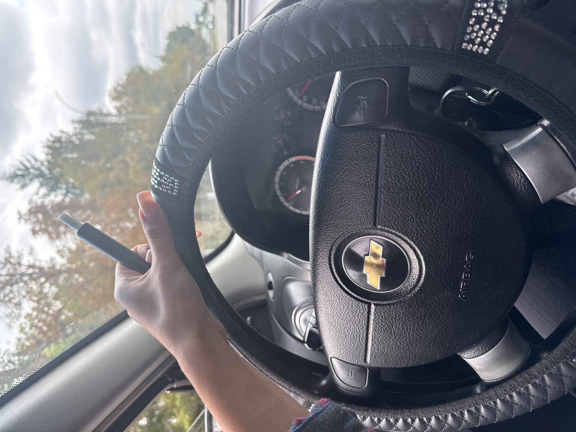 A person's left hand rests on the steering wheel of a car. The steering wheel is black, has a textured grip, and is decorated with rhinestones. The car's dashboard is visible behind the steering wheel, including the speedometer and other gauges. The image appears to be taken from inside the car, looking forward through the windshield. Outside the car, trees and a cloudy sky are visible, suggesting an outdoor, possibly suburban or rural setting. The scene does not depict any specific location in Basarabeasca, Moldova.
