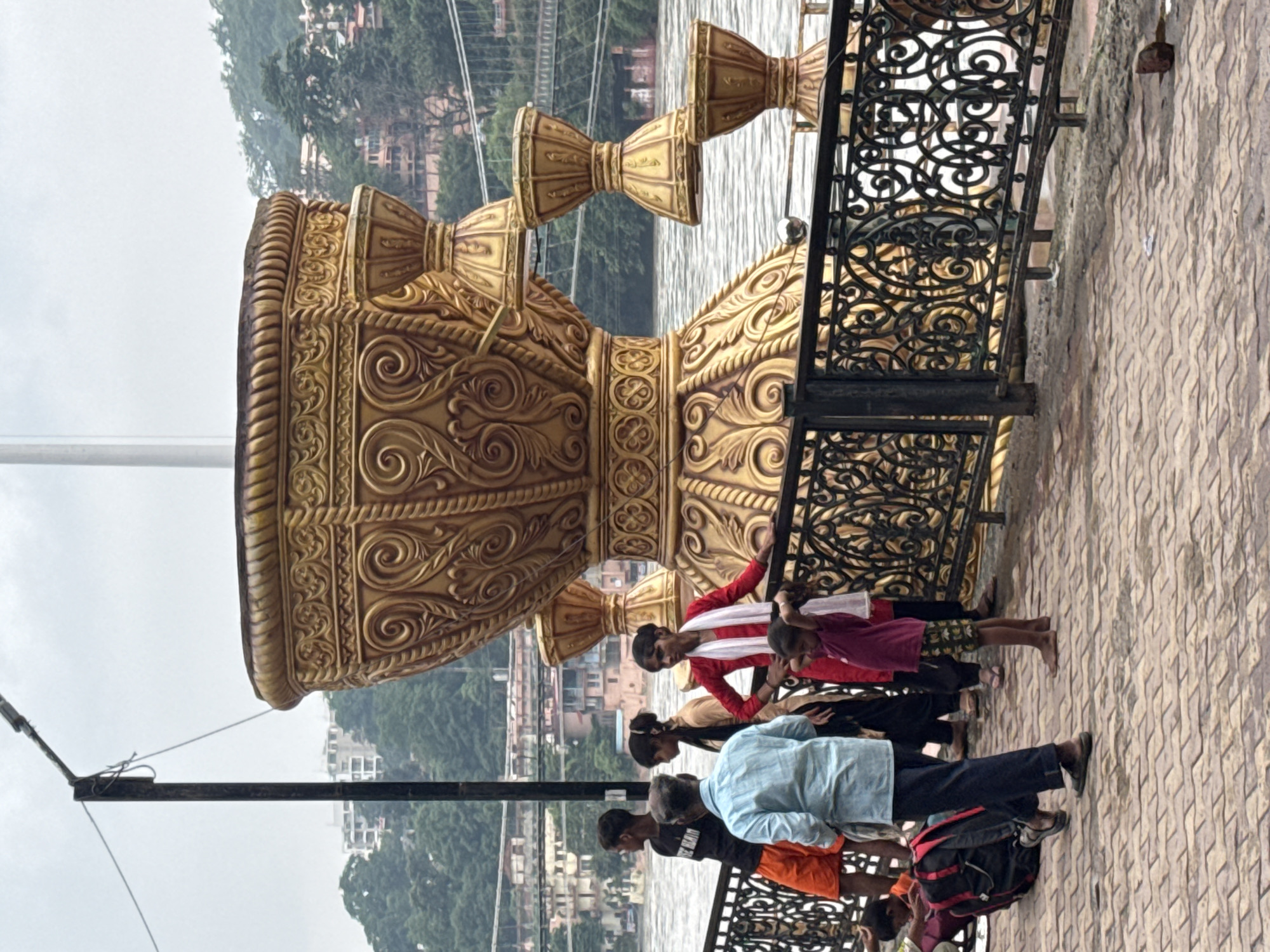 A group of people stand near a large golden sculpture by a river in Rishikesh, India. The sculpture resembles a giant, ornate vase with several smaller bell-shaped elements attached. The people, a mix of adults and children, are dressed in casual clothing. One woman in a red outfit and white scarf stands near the sculpture, while a young child in a purple shirt and patterned shorts stands barefoot. To the left, a man in a light blue shirt and dark pants walks away from the camera, and a young boy in orange shorts and a black t-shirt stands looking towards the river. In the background, the wide, flowing river is visible, with a suspension bridge crossing it further in the distance. Lush green hills and buildings can be seen on the far bank of the river. The foreground consists of a brick-patterned walkway and a decorative black metal railing. The sky is overcast.