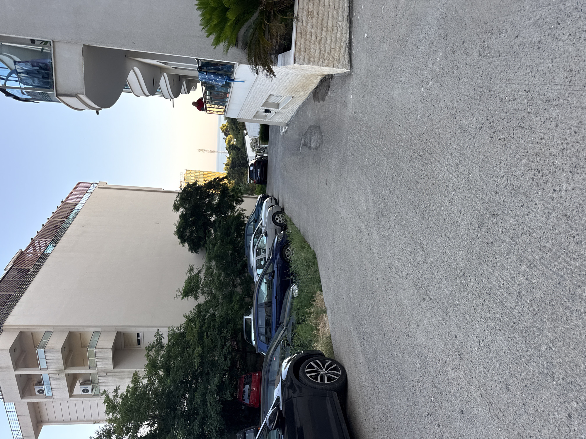 The image shows a street with cars parked on the side, in front of buildings. The street is paved and has some green areas with trees and grass along the side. In the background, there are buildings, some of which appear to be residential or hotels, with balconies and windows. There is also a view of the sea and the sky. In the foreground, there are several cars parked on the asphalt. One person, wearing a red shirt, is standing on a balcony in the upper right portion of the image. The overall scene depicts a typical urban or semi-urban environment in a coastal area. The location is identified as Boreti, Montenegro, which is consistent with the presence of the sea and architecture.