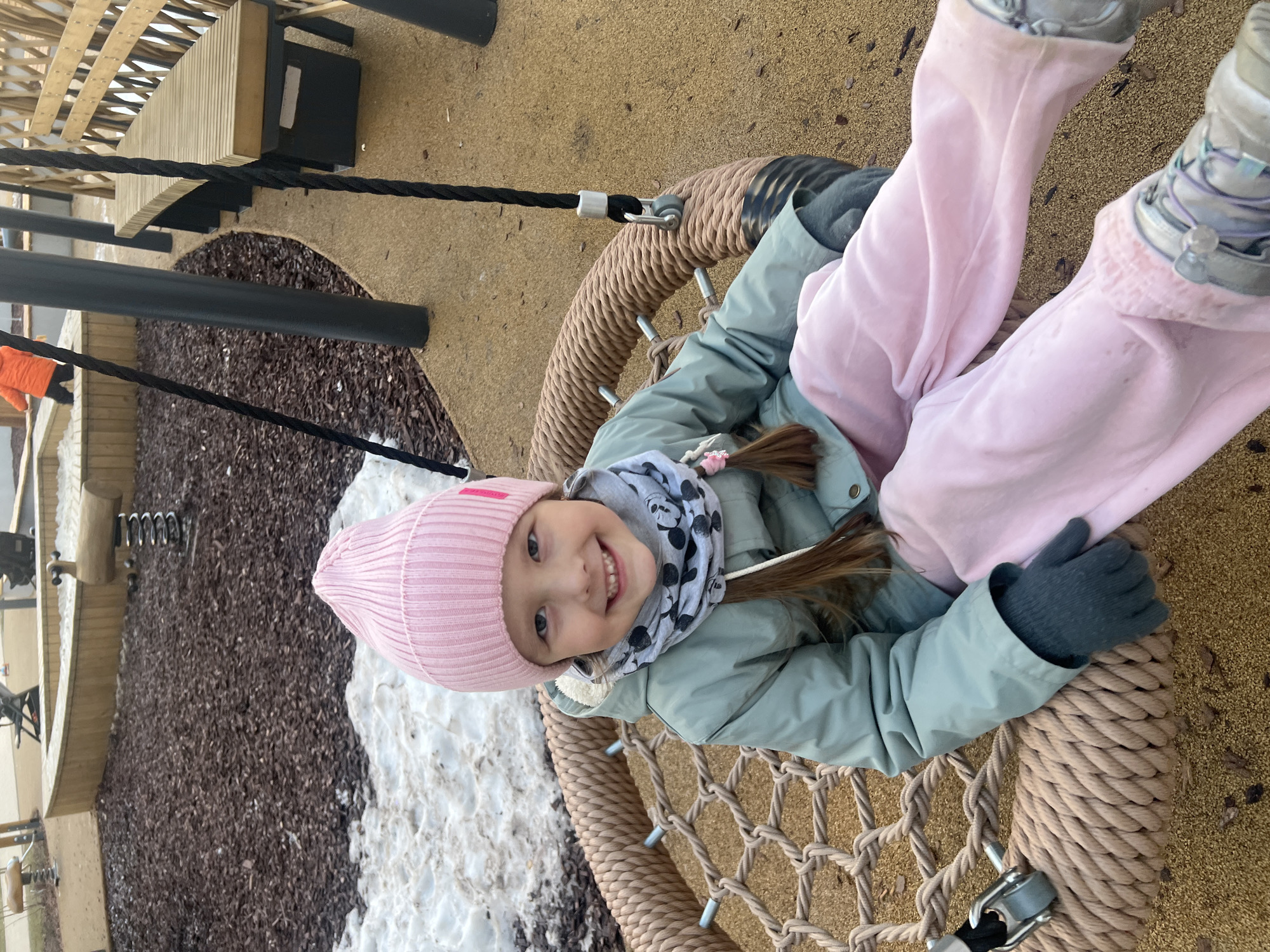 A young girl sits in a circular rope swing, smiling at the camera. She wears a pink knit hat, a light gray jacket, a gray and white patterned scarf, gray gloves, pink pants, and silver boots. The swing hangs from black ropes. The surrounding ground includes areas of brown mulch and patches of snow. The scene appears to be a playground or recreational area. Elements of wooden structures are visible in the background. The city of Chemoshur, Russia is not apparent in this image.