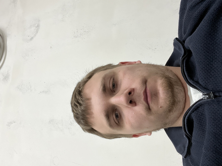 A close-up portrait of a fair-skinned man with short blonde hair and a light beard. He looks directly at the camera with a neutral expression. He is wearing a dark blue zip-up hooded sweatshirt. The background is a plain white wall with some discoloration and markings. The image is rotated slightly to the right.