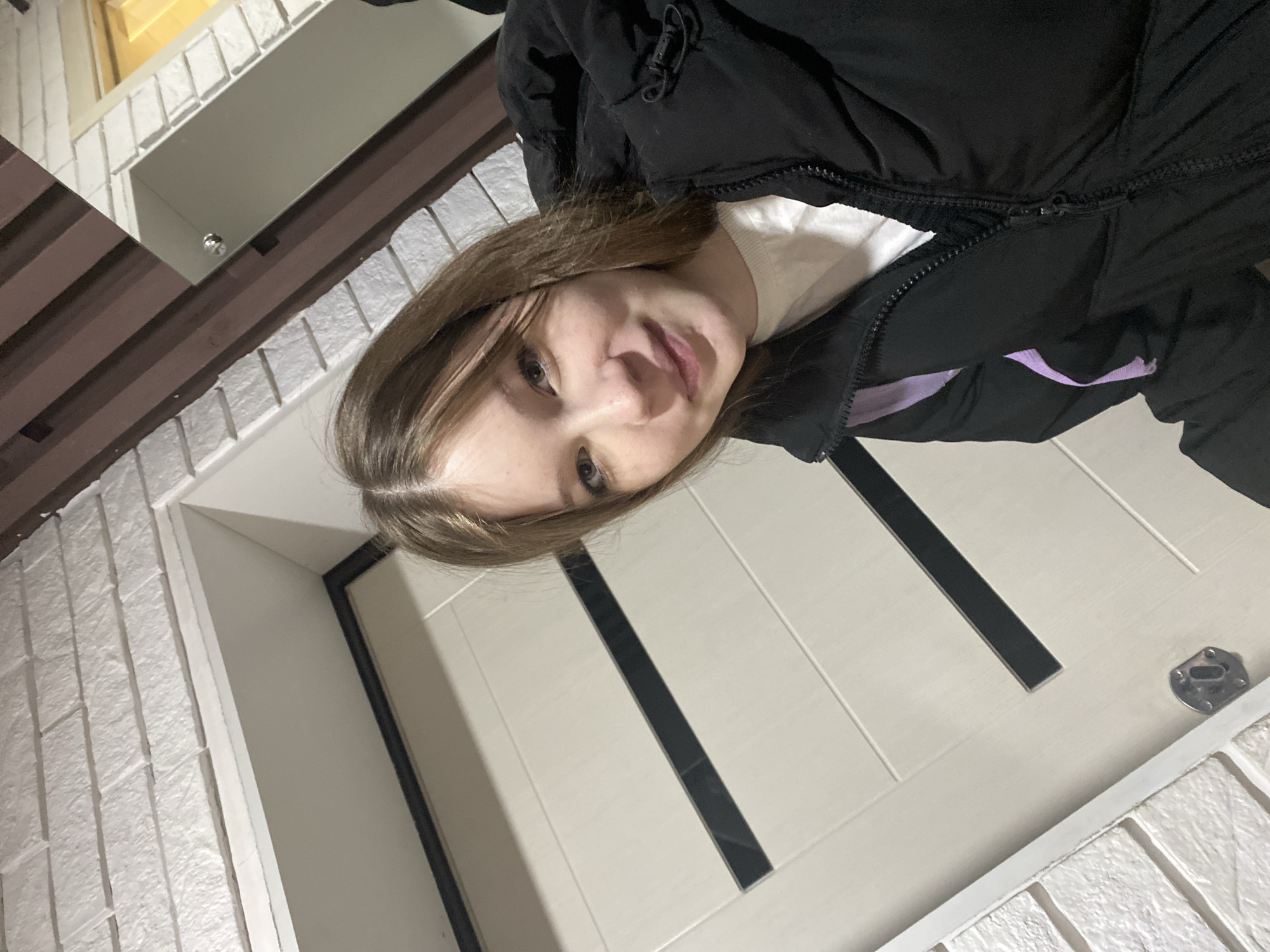 A person wearing a black down jacket is standing in front of a white door with black vertical inlays. The location appears to be Chaikovsky, Russia, based on architectural styles. The person is facing the camera. The surrounding structure includes textured white brick and brown wooden trim.