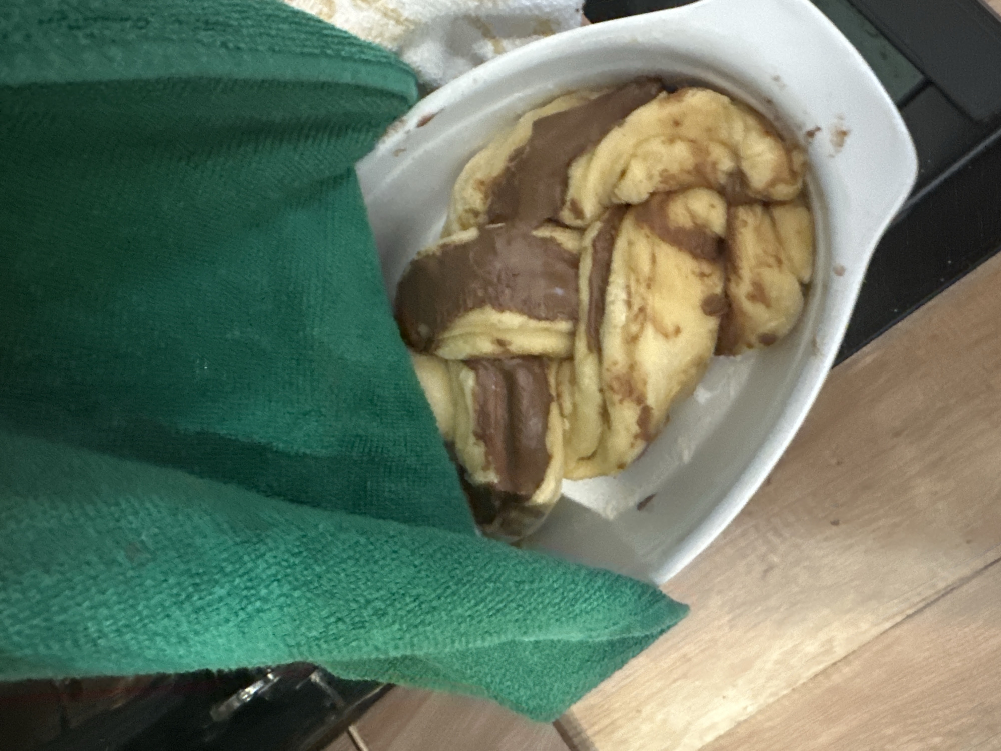 A dish of baked goods with a chocolate topping sits partially covered by a green towel. The dish is white and angular, placed on a wooden surface. To the right is a black surface.
The location is not determinable from the image.