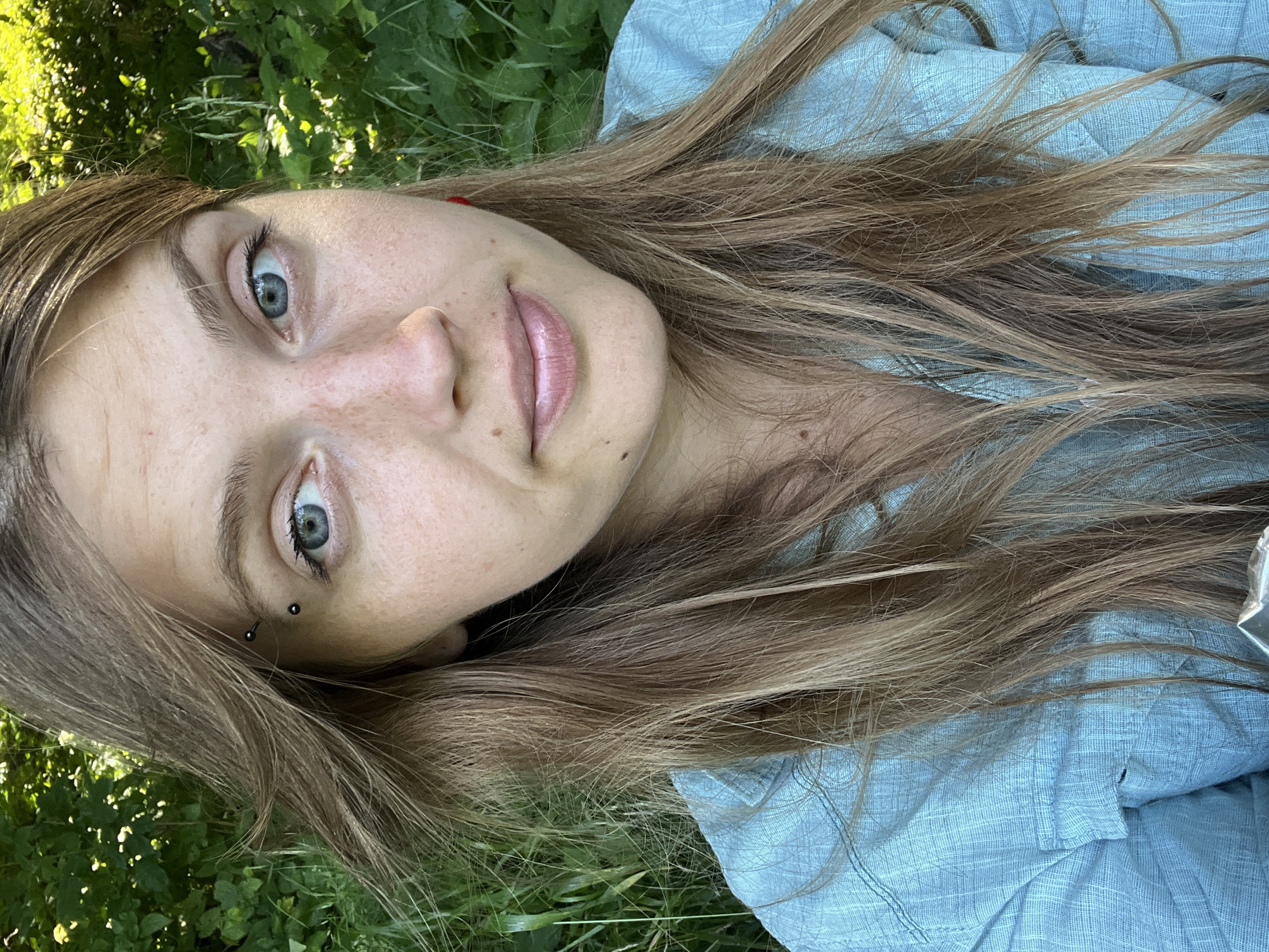 A young woman with light brown hair and blue eyes is pictured from the chest up, outdoors. She has a piercing above her left eyebrow and freckles across her nose and cheeks. She is wearing a light blue collared shirt. The background is filled with lush green foliage. The location is in the village of Garmonavichy, in the Garmanavichy rural council of Belarus.