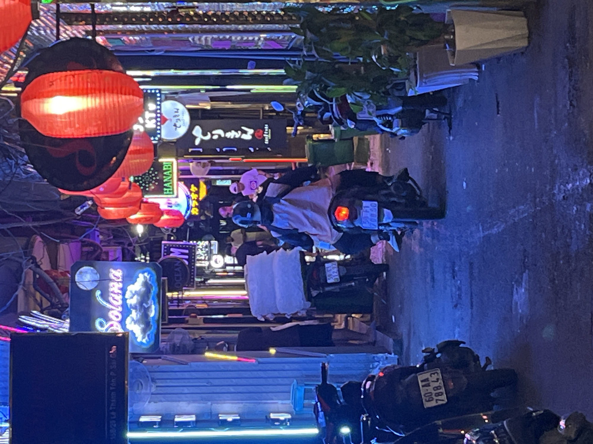 The image depicts a narrow street scene at night, likely in Thủ Đức, Vietnam, characterized by bright, colorful neon lights and signage. In the foreground, several motorcycles are parked. One motorcycle on the left has a license plate that reads "60-AA 788.43". A person on a motorcycle, wearing a helmet and a jacket, is seen from behind, navigating down the street. This motorcycle has a large, light-colored load strapped to its rear. Further down the street, other individuals are visible, some standing and others on motorcycles. The buildings lining the street are adorned with various signs, including "Solana" with a cloud graphic, and what appear to be Japanese characters and other commercial signage illuminated with neon and LED lights. Red lanterns are hung overhead, adding to the vibrant nighttime atmosphere. The street surface appears wet in places, suggesting recent rain or cleaning. The overall impression is a bustling urban night market or entertainment district.