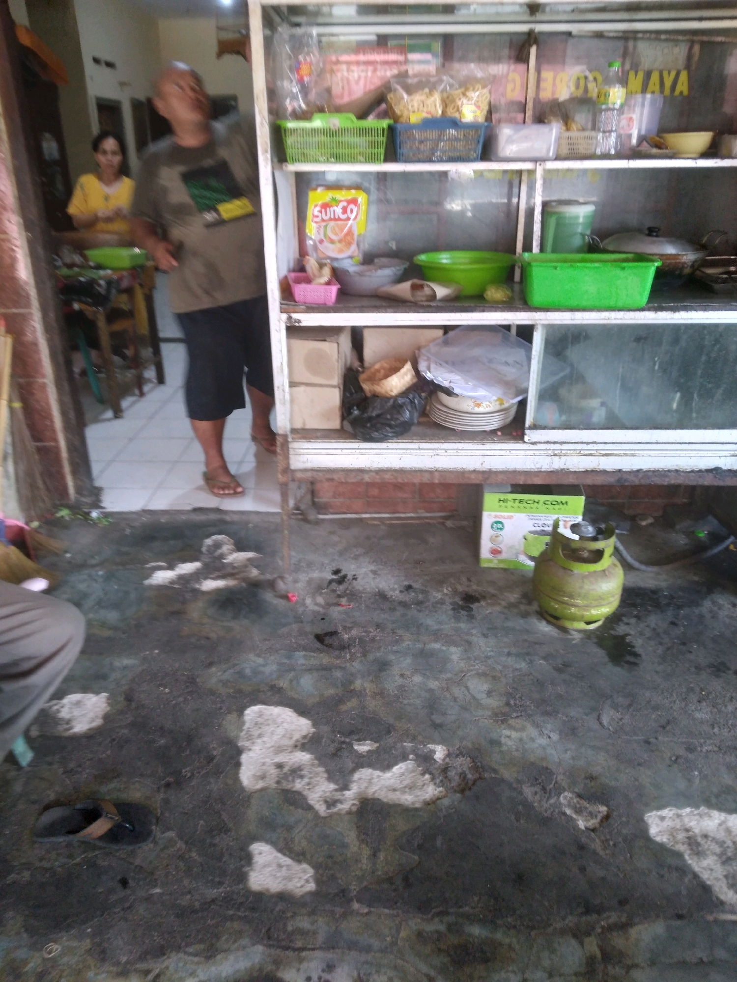 A man and a woman stand inside a food stall in Purwokerto, Indonesia. The man, wearing a brown t-shirt and black shorts, looks up and to his right, his left hand extended. The woman, in a yellow t-shirt, stands behind him. The stall features shelves stocked with various food items and kitchenware. A gas cylinder sits on the floor in front of the stall. The ground outside the stall is wet and dirty, with a pair of flip-flops visible. The overall scene depicts a busy, everyday scene at a local eatery.