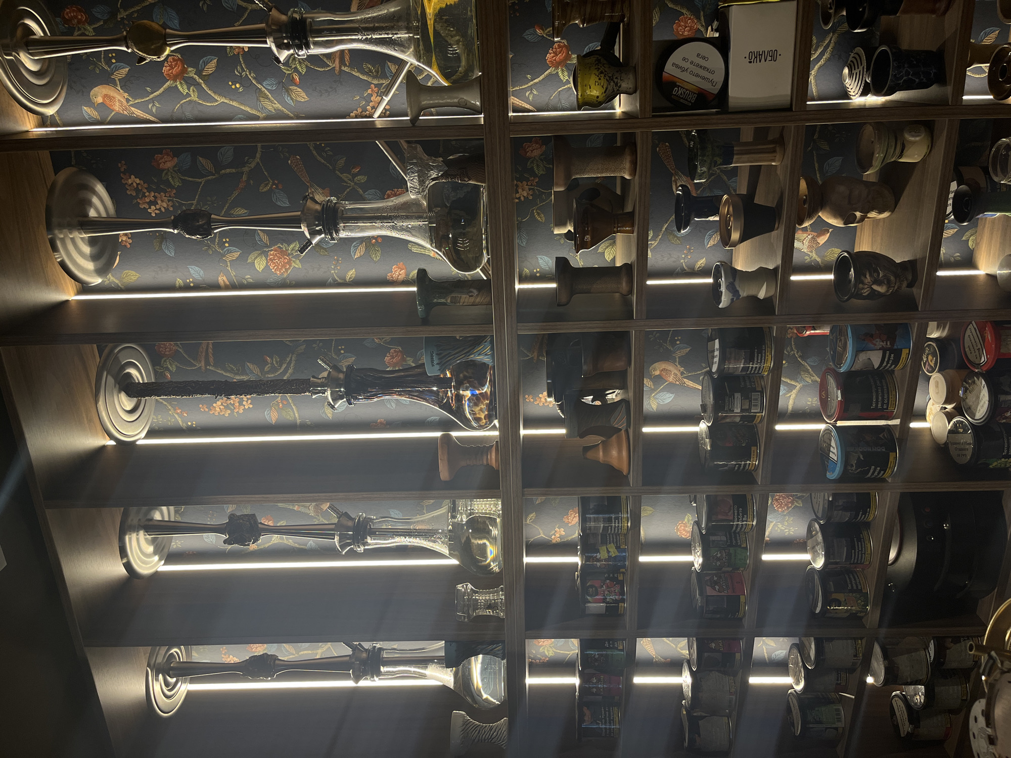 The image shows a well-lit shelving unit filled with hookahs and related accessories. The shelves are made of wood, and vertical strip lights illuminate the items. The background wallpaper features a dark blue floral pattern with birds. On the top shelves, several elaborate hookahs with glass bases and metallic stems are displayed. The middle shelves are populated with various hookah bowls, some made of ceramic and others of clay, in different shapes and colors. The bottom shelves contain numerous cylindrical containers, likely tobacco or shisha flavors, along with some additional hookah accessories. The overall presentation suggests a retail environment, possibly a hookah lounge or a specialized shop. The scene is static, with no people or immediate actions depicted. This establishment is located on Hristo Botev Street in Plovdiv, Bulgaria.