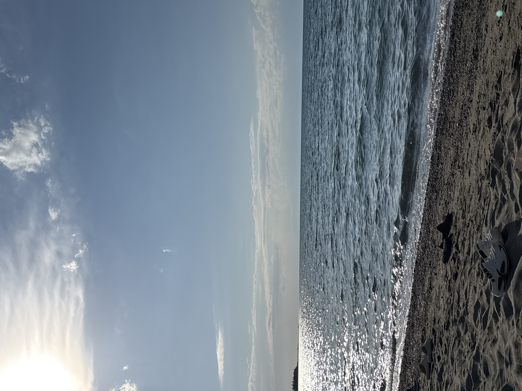 This is a photograph taken at Issyk-Kul Lake in the city of Balykchy, Kyrgyzstan. The image captures a serene beach scene with a sandy shore in the foreground and the expansive, shimmering waters of the lake stretching towards the horizon. A bright sun is visible in the upper left corner, casting a luminous glow on the water. Several pairs of footwear, including white athletic shoes and dark sandals, are scattered on the sand near the water's edge. The sky is a clear blue, dotted with wispy white clouds, suggesting fair weather. The overall atmosphere is one of tranquility and a peaceful day at the lake.