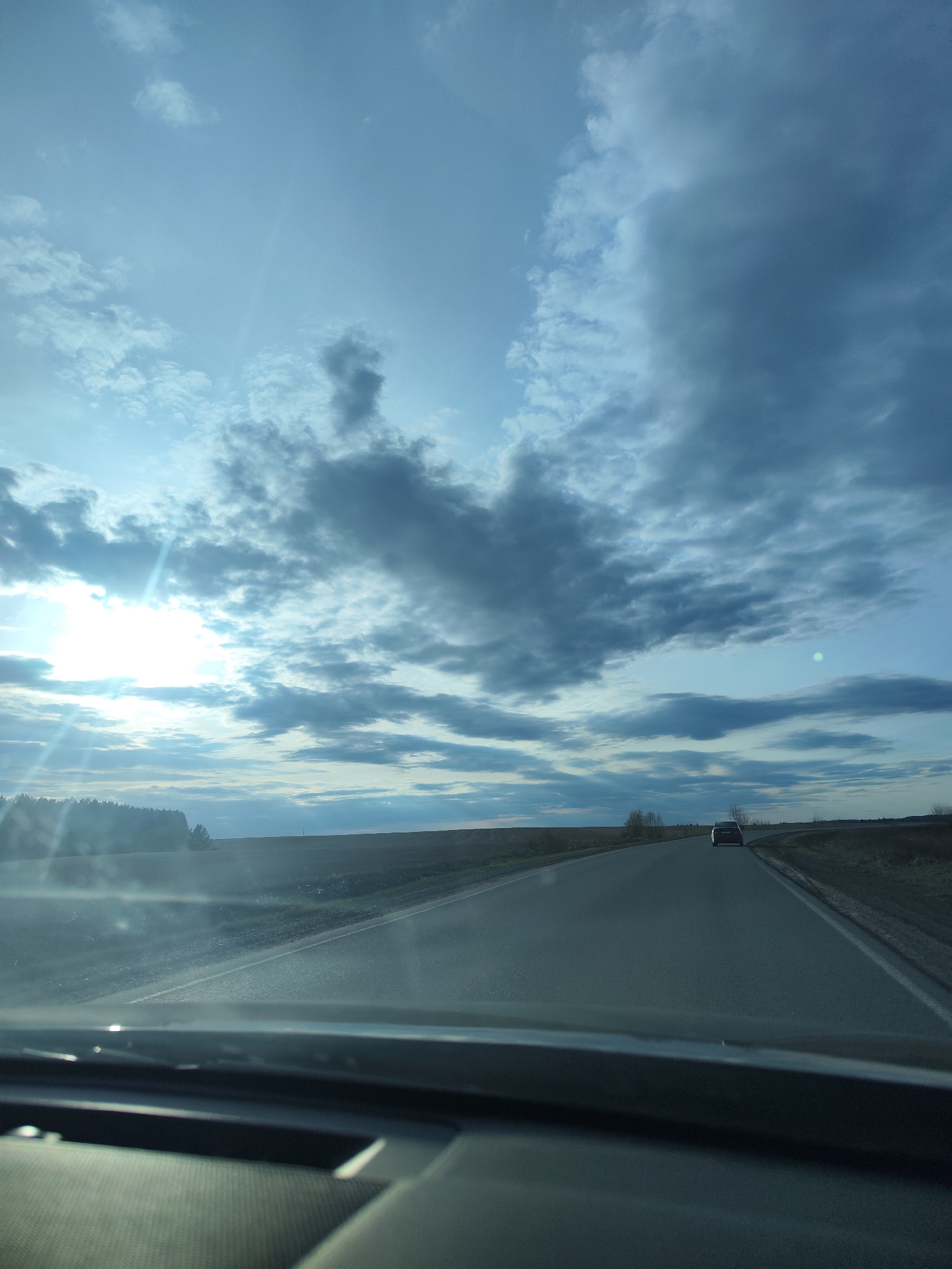The image depicts a view from inside a vehicle, likely a car, driving on a rural highway. The sky occupies a large portion of the frame, showcasing a mix of blue sky and scattered cumulus clouds, with the sun appearing as a bright glare on the left side. The landscape consists of flat, open fields on either side of the road, with a sparse treeline visible in the distance to the left. In the distance, a dark-colored car is seen traveling in the same direction. The road ahead appears straight and flat. The interior of the car is visible in the lower portion of the image, showing the dashboard and part of the steering wheel.