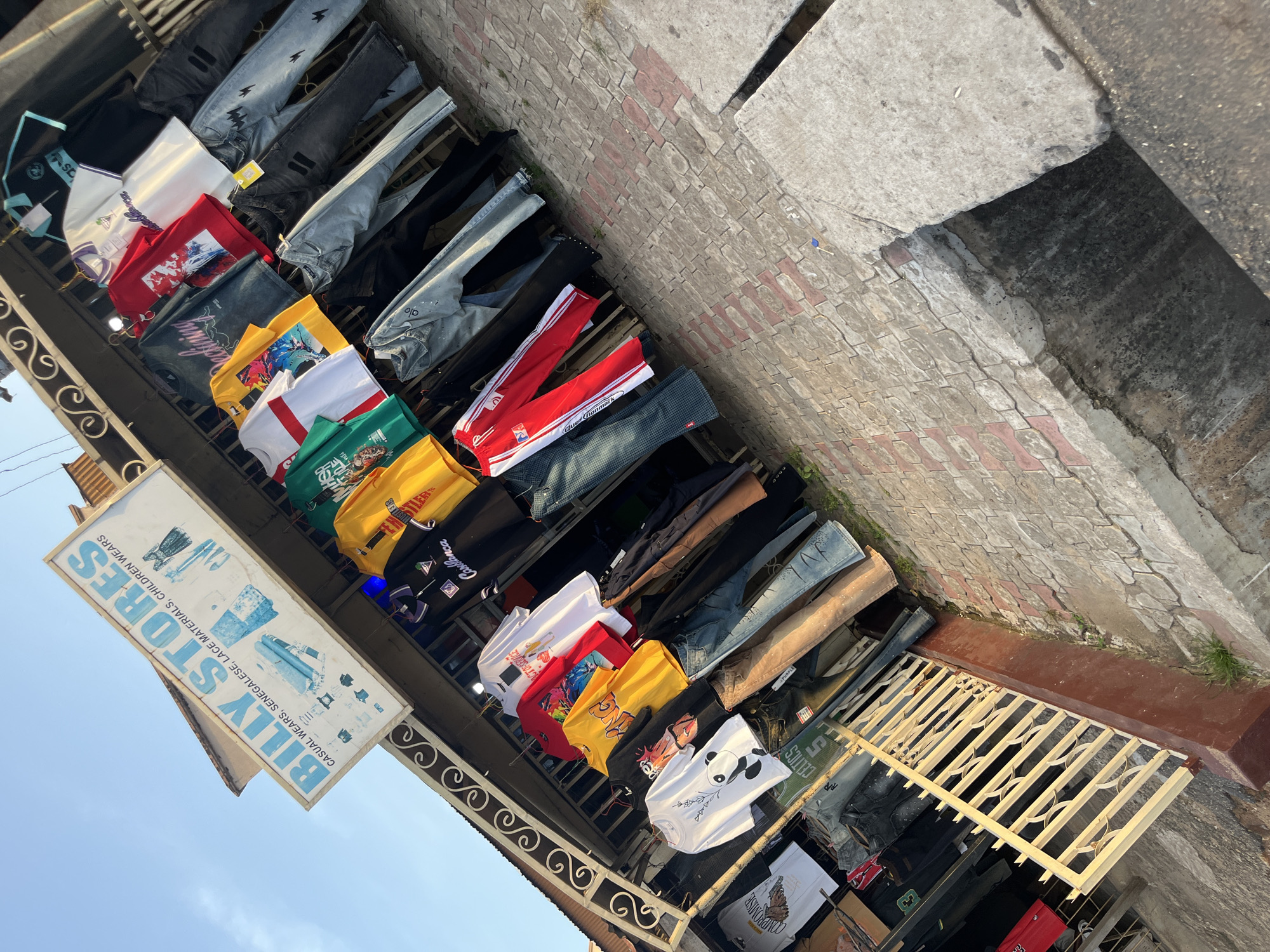A street market stall selling clothing is depicted in the image. A sign with the text "BILLY STORES CASUAL WEARS, SENEGALESE, LACE MATERIALS, CHILDREN WEARS" is prominently displayed. A variety of apparel, including t-shirts and jeans, are hung on display racks. The stall is set up along a brick-paved sidewalk, with a drainage ditch visible to the right. The sky above is clear and blue.