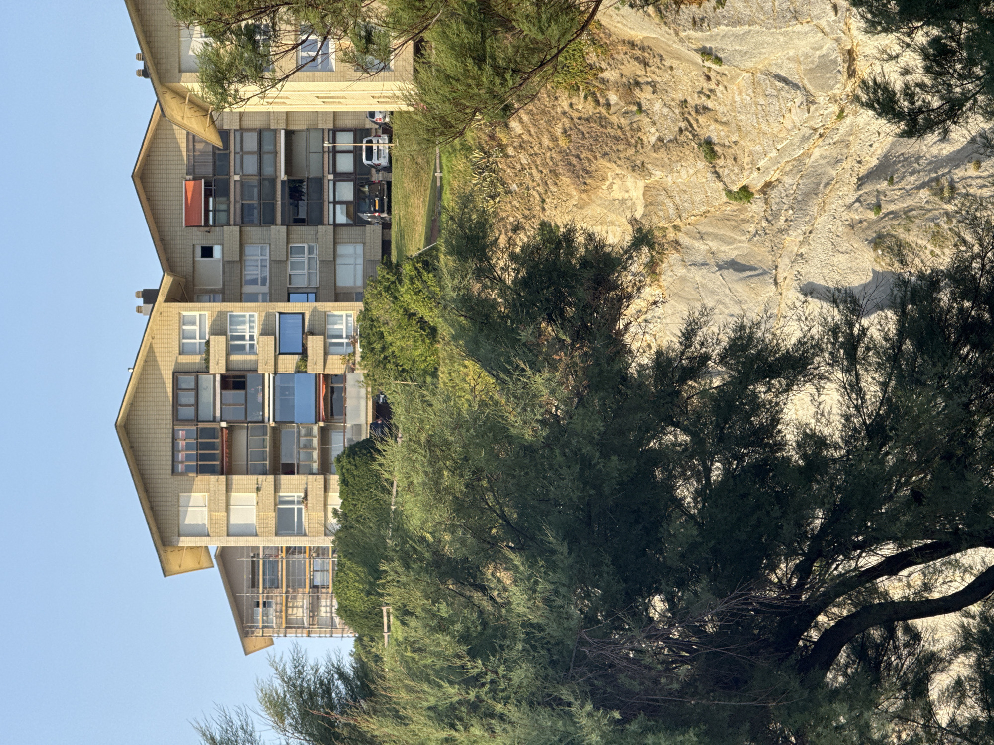 The image depicts a residential building complex situated on a cliffside in Sopela, Spain. The buildings are multi-story structures with light-colored brick facades and distinctive gable roofs. Several apartments feature balconies with glass railings and some have awnings, including an orange one visible on the central building. The buildings are surrounded by lush green vegetation, including trees and bushes, which cascade down the cliff. The cliff face itself is a prominent feature, showing layered rock formations in shades of beige and tan, with sparse green growth. A few vehicles, including vans and a car, are parked on the grass area in front of the buildings. The sky above is a clear, light blue.
