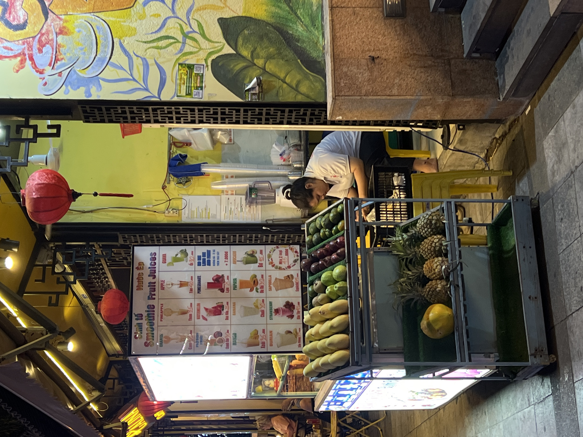 Here's a detailed description of the image:
**Setting:** A street scene in Nha Trang Ward, Vietnam. The setting appears to be in an older part of the city, based on architectural features.
**Subjects:** A woman wearing a white shirt and dark shorts is working in a small fruit stall. She's bent over, possibly preparing fruit or organizing the stall.
**Objects:**
* **Fruit Stall:** The stall is prominently featured, with a variety of fruits displayed, including pineapples, mangoes, avocados, pomegranates, apples, and other tropical fruits. A menu board lists different smoothie and juice options with prices.
* **Menu Boards:** There are brightly lit signs displaying the menu and prices for the smoothie and juice offerings.
* **Red Lanterns:** Traditional red lanterns hang near the stall, likely adding to the ambiance, especially in the evening.
* **Seating:** A yellow plastic stool.
* **Building Elements:** The backdrop includes parts of buildings – a wall with a painted mural featuring foliage, and another structure with steps.
**Actions:** The woman is actively engaged in tasks related to the fruit stall business.
**Environment:** The scene is at dusk or night, illuminated by artificial lighting from the stall.
