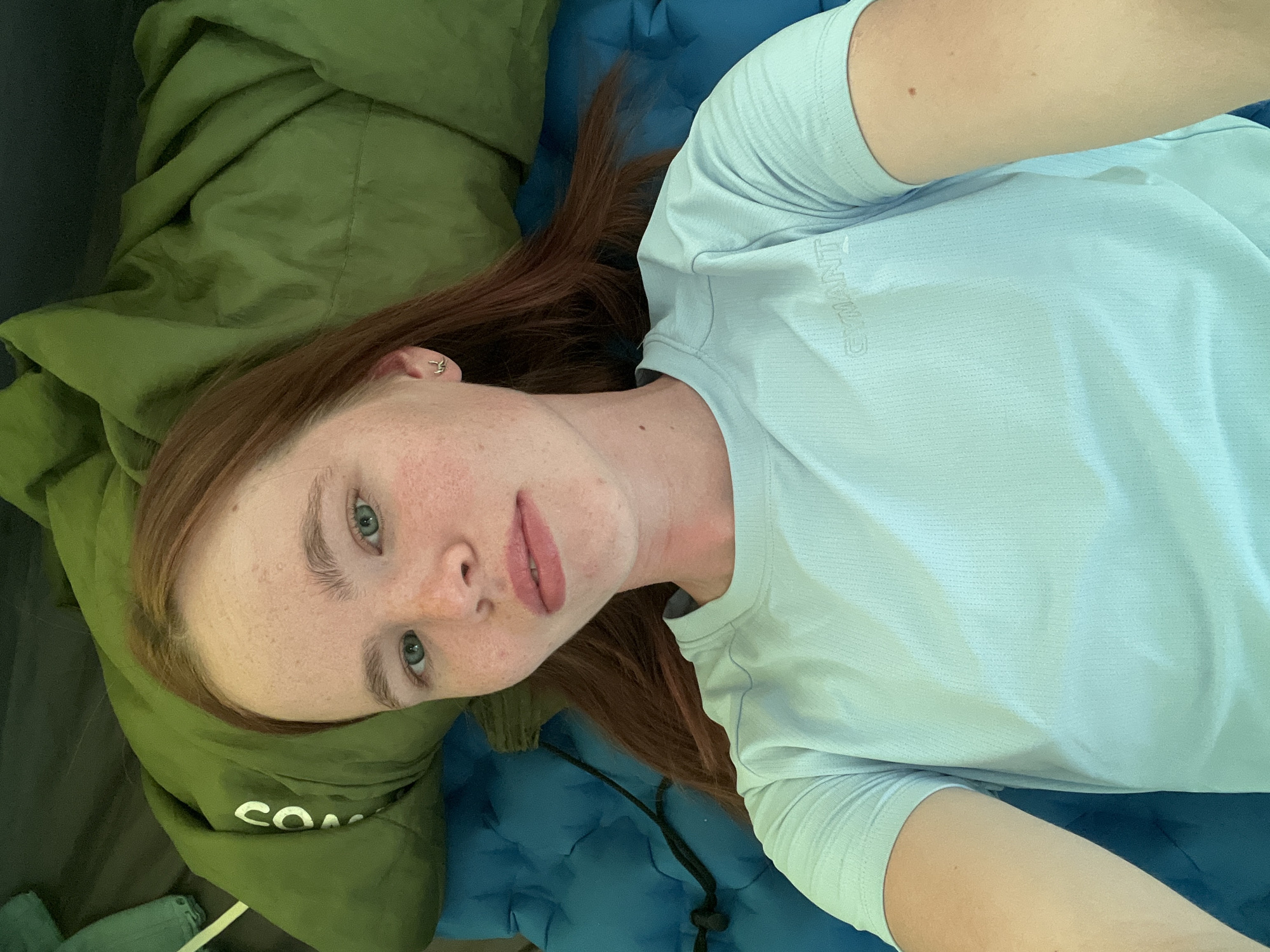 A fair-skinned person with red hair and freckles lies on a blue sleeping pad inside what appears to be a green tent. The person is wearing a light blue t-shirt. The location is unknown and there is no evidence to suggest it is in Khvalynsk, Russia.