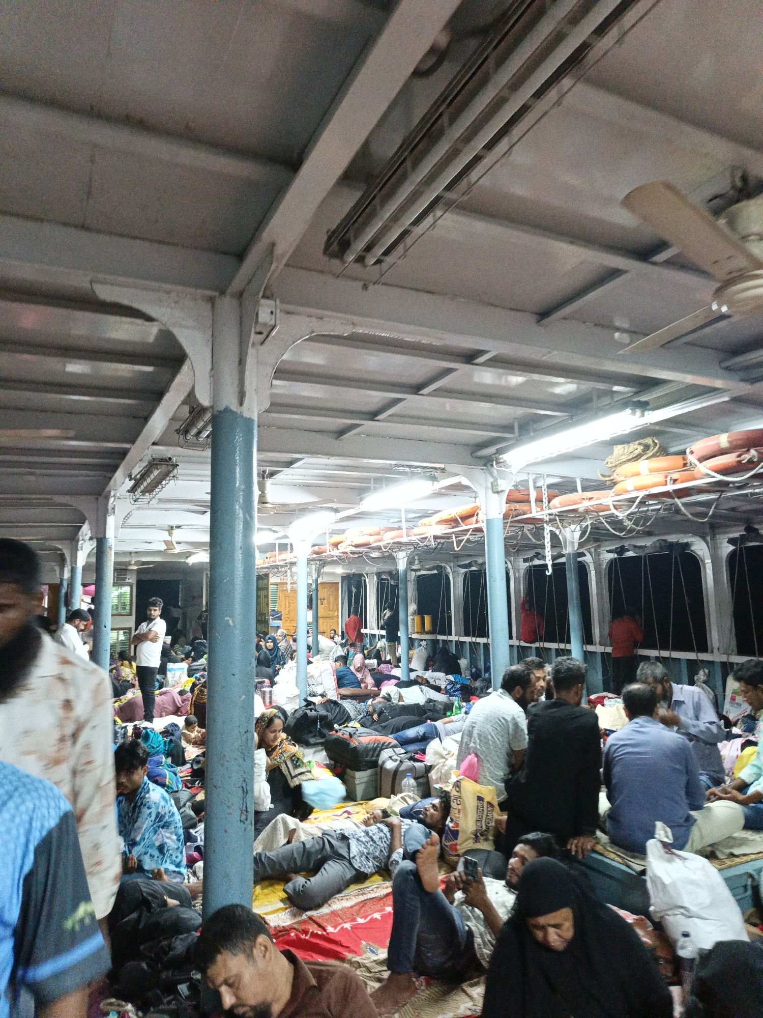 The image depicts the interior of a ferry in Goshairhat, Bangladesh, filled with people. The ferry's structure is visible, including metal beams, columns, a ceiling with fluorescent lights, and ceiling fans. Along the sides, life preservers are secured. Passengers are densely packed on the floor, many lying on mats or blankets. Some are sitting, and a few are standing. The scene appears crowded and possibly chaotic. Passengers are clothed in a variety of traditional and modern attire. Bags and personal belongings are scattered among them. Some people are interacting, while others are resting or looking at their phones.