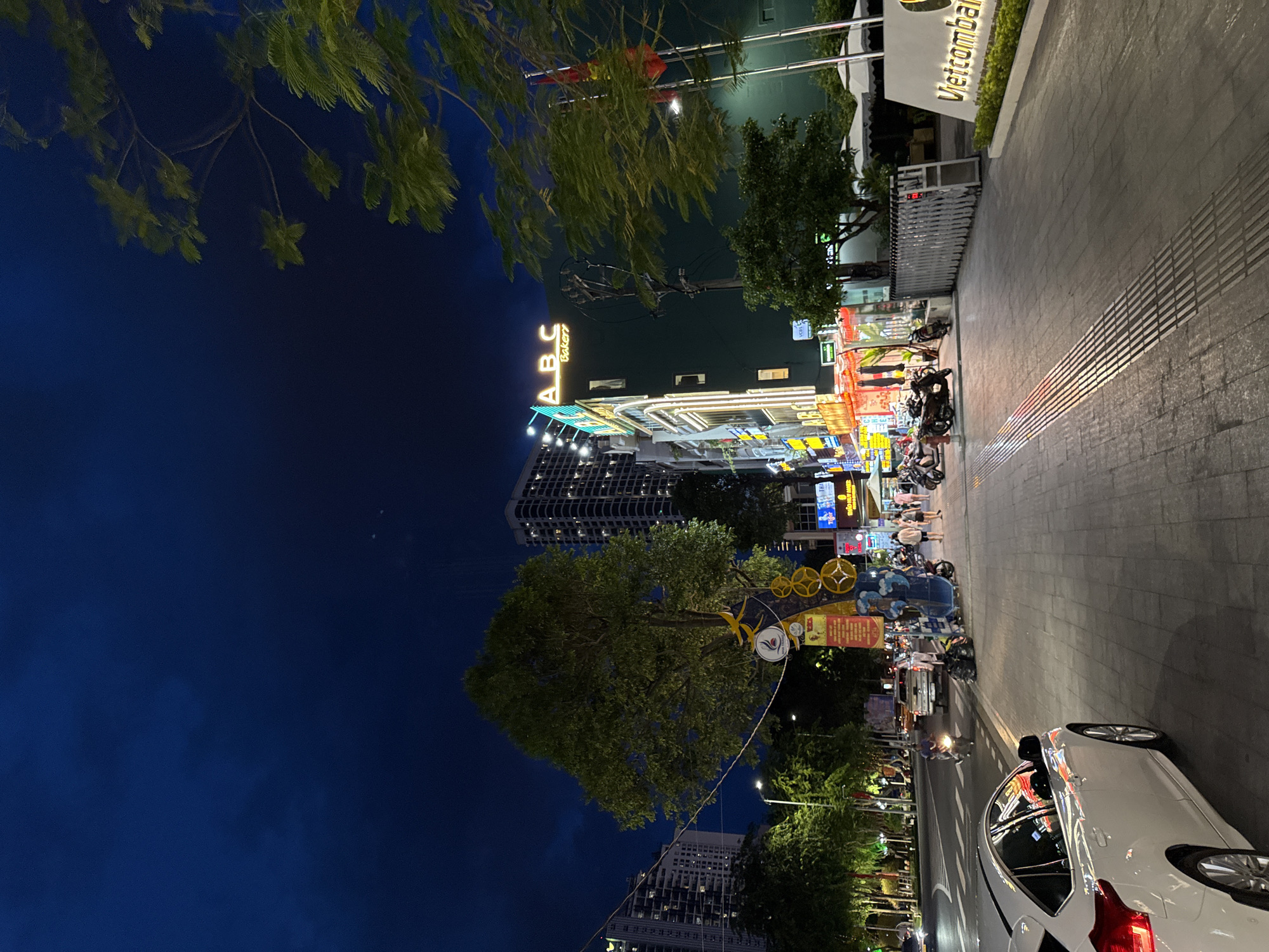 The image shows a street scene at night in Nha Trang, Vietnam, near the ABC Bakery. A white car is parked on the left side of the street, facing away from the camera. The street is paved with large tiles, and there are trees and lampposts along the sidewalk. Several motorcycles are parked on the sidewalk, and a few people are walking by. Buildings line the street, with neon signs and lights illuminating the scene. One building has a large "ABC Bakery" sign with neon lights. A white sign with green text reads "Vietcomban...". The sky is dark blue, indicating it is nighttime.