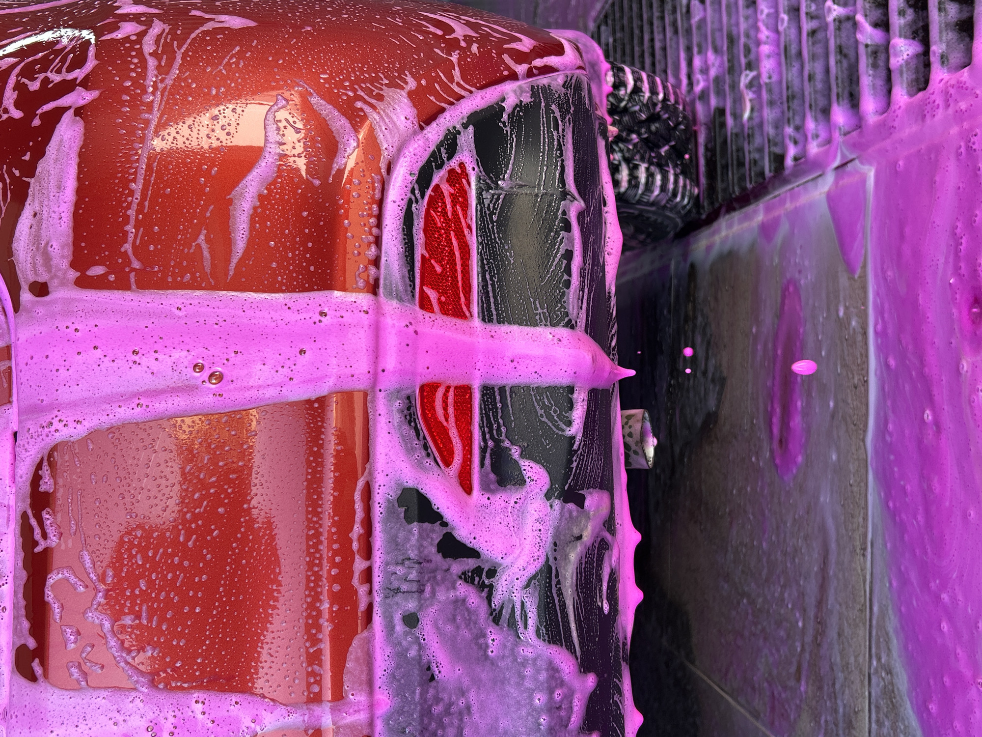 The image depicts the back end of a red car undergoing a wash at what appears to be an automated car wash facility. Pink soapy foam covers the car's body, rear light, bumper, and nearby surfaces, indicating the use of a colored soap solution for cleaning. The car's tire and the surrounding structures of the car wash are partially visible in the background. The location is presumed to be in Chaikovsky, Russia.