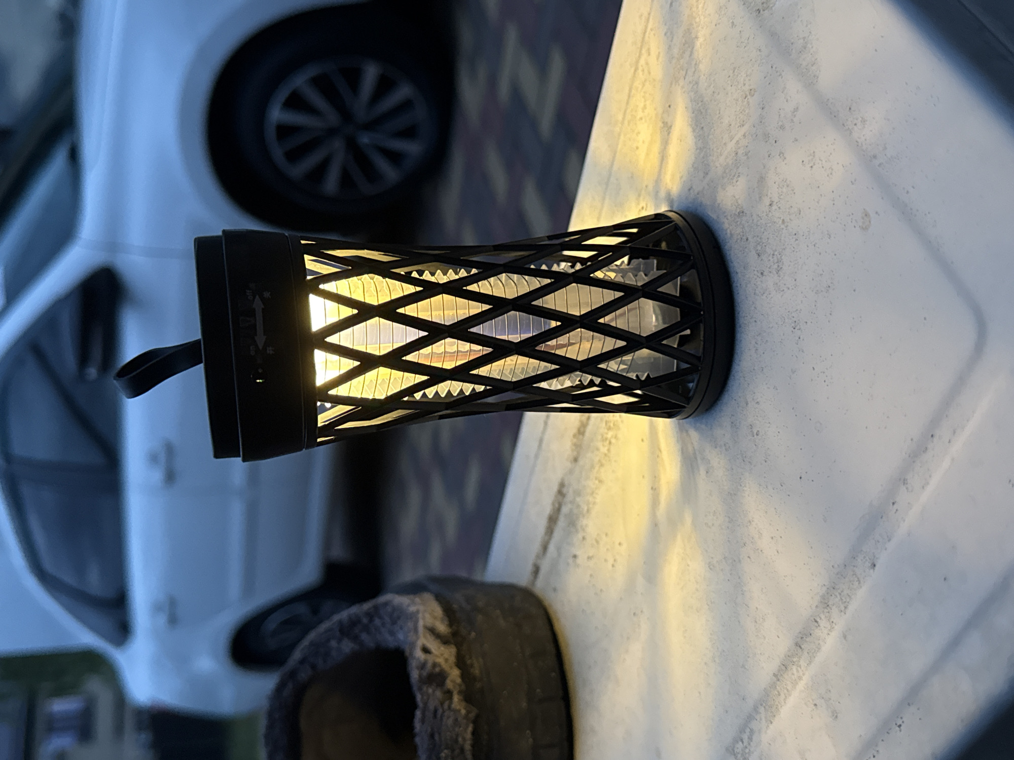 A black, decorative lantern with an intricate, open-work diamond pattern casing illuminates a white, textured surface in a low-light outdoor setting. The lantern emits a warm, yellow-orange glow from an internal light source. Attached to the side of the lantern is a black strap or handle with the word "OUTDOOR" faintly visible. On the lantern's body, near a slider switch, are symbols including a plus sign, a minus sign, and Chinese characters "开" (on) and "关" (off) with a double-headed arrow, indicating controls for the light.
In the blurred background, a white car with a dark, spoked wheel is partially visible. A dark, fuzzy or textured object rests below the lantern in the foreground. The ambient light suggests it is dusk or nighttime. No people are present.