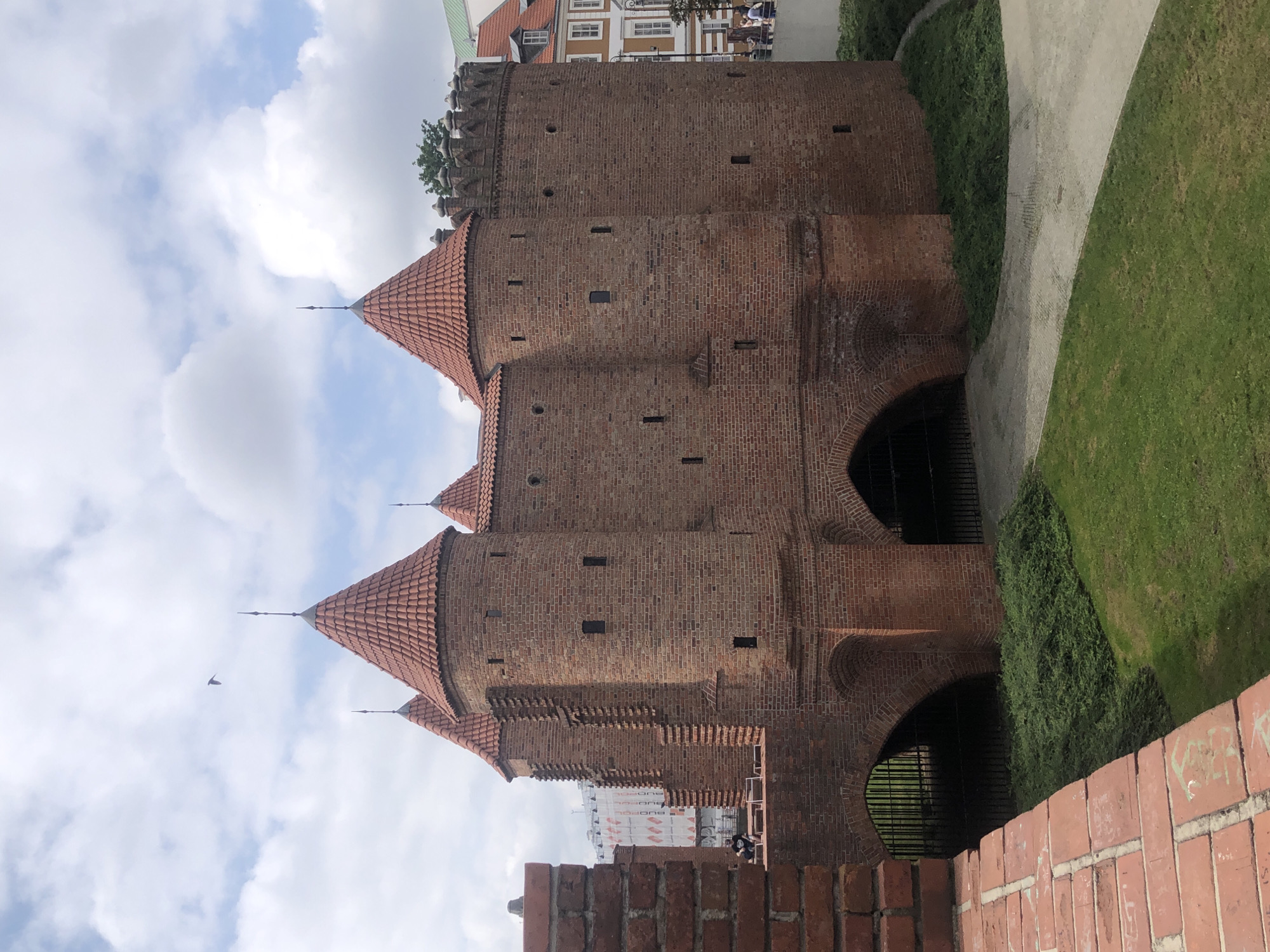 The image shows the Warsaw Barbican, a historic fortification in Warsaw, Poland. The Barbican is constructed of red brick and features multiple round towers with conical tiled roofs. An arched gateway with a metal grate is visible, leading into the structure. In the foreground, a brick wall with graffiti is present, and to the right, a path winds through a grassy area. The sky is partly cloudy. In the background, modern buildings and some trees are visible. A bird is flying in the sky above the Barbican. A few people are seen near the Barbican and on a walkway to the right.