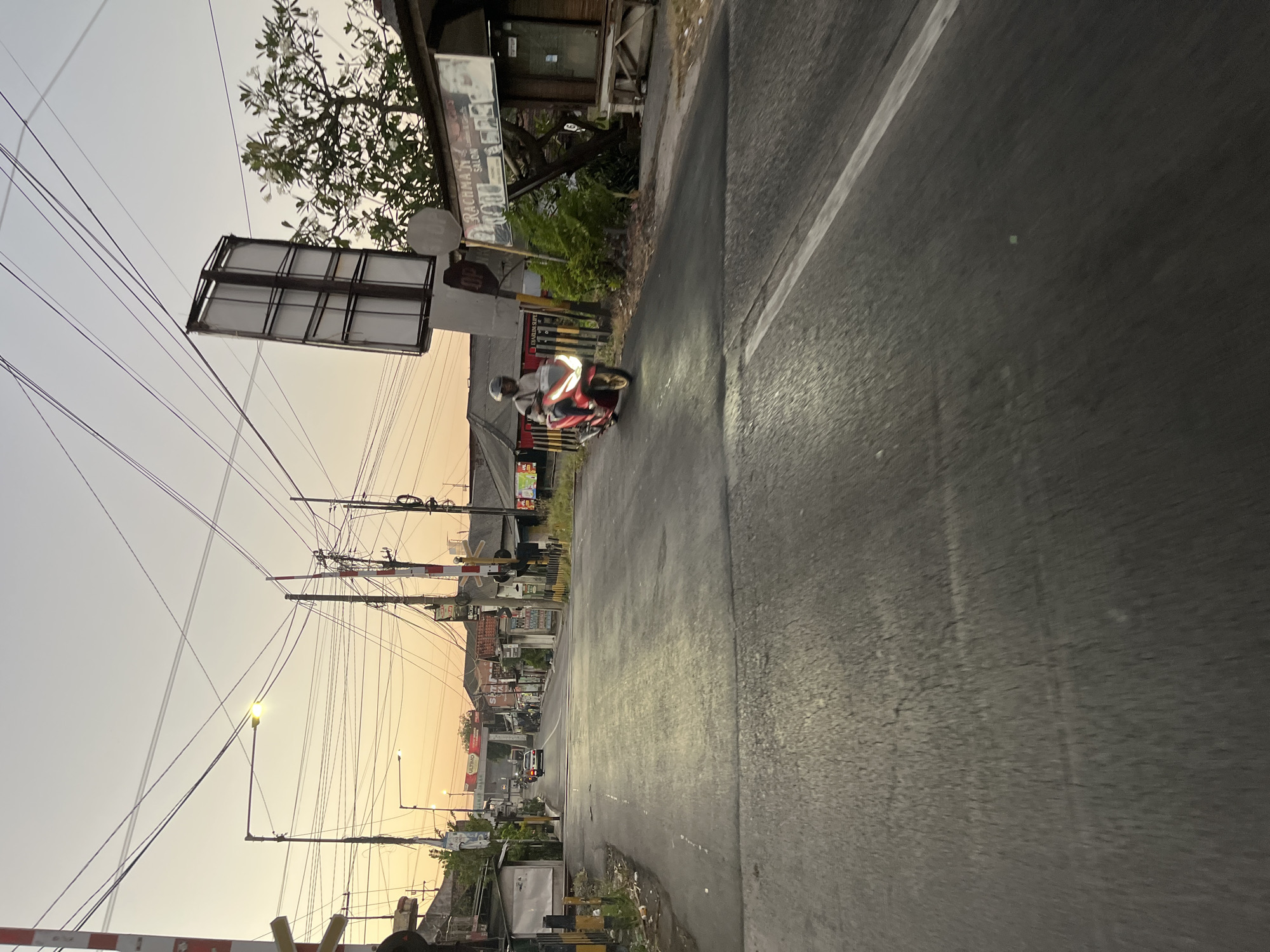 The image depicts a street scene in Boro, Indonesia, at dusk. A red motorcycle with a rider wearing a helmet is moving away from the camera on the right side of the road. The road is paved and has a white line marking the lanes. There are various buildings and structures lining the street, including shops, houses, and utility poles with numerous wires. A railroad crossing gate is visible on the left side of the road. The sky is a pale orange and yellow, indicating the sunset. The overall impression is a busy street in a residential or commercial area during the transition from day to night.