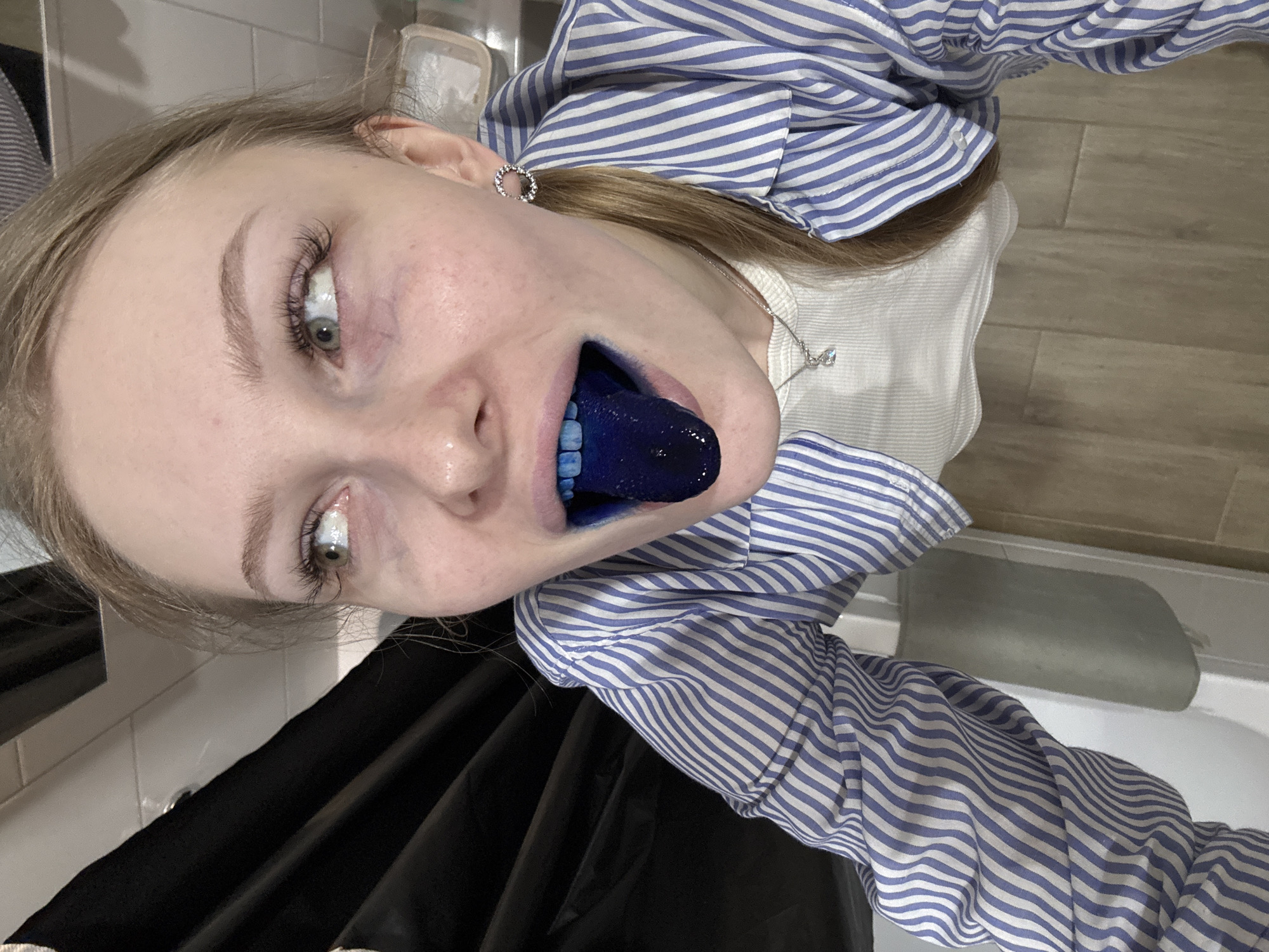 The image is a vertical selfie of a young person, likely female, with light hair and fair skin, taken indoors, possibly in a bathroom. The subject's head is tilted to the right, and they are looking directly at the camera with wide, light-colored eyes. Their mouth is open, revealing a strikingly vibrant, solid blue tongue, suggesting consumption of a blue-dyed food or drink.
The person is wearing a blue and white striped button-up shirt over a white ribbed top. A delicate silver necklace with a small, clear pendant hangs around their neck, and a silver hoop earring is visible in their left ear. Their expression appears playful or surprised, drawing attention to their unusually colored tongue.
The background includes light-colored rectangular wall tiles on the left side of the frame and a light-toned wooden or wood-effect tiled floor on the right. A white fixture, possibly the rim of a sink or bathtub, is visible in the bottom right corner, reinforcing the indoor, bathroom-like setting. The lighting is artificial, characteristic of an indoor environment. There is no visible text in the image.