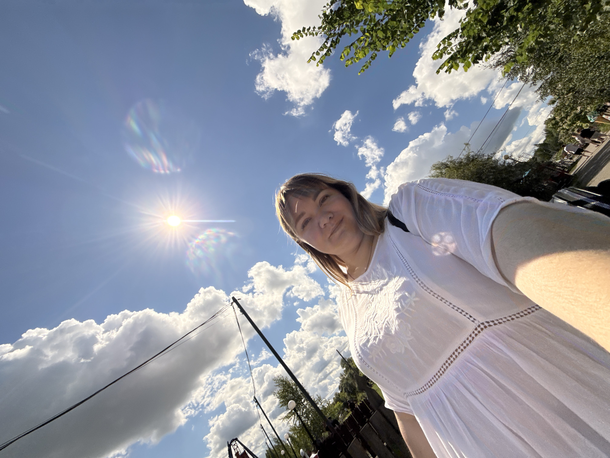 The image is a low-angle, wide-shot, selfie taken outdoors in daylight, possibly in the city of Kotovo, Russia. The sky dominates the frame, filled with a bright blue expanse and numerous fluffy white clouds. A strong sun with visible rays and lens flare is positioned in the upper center of the image.
In the foreground, a person is visible from the chest up, facing the camera. They have blonde hair and are wearing a white, flowing dress with embroidered details around the neckline and torso. Their right arm is extended, holding the camera.
The background reveals an outdoor environment with several elements. To the left, there is a dark wooden fence and some greenery, including trees. Some street lights or decorative lights on poles are also visible. Further into the background, a pathway or street can be seen with several people walking away from the camera. Trees and more foliage line the sides of the pathway. Overhead, power lines are strung between poles. The overall scene suggests a casual outdoor setting, possibly a park, street, or event.