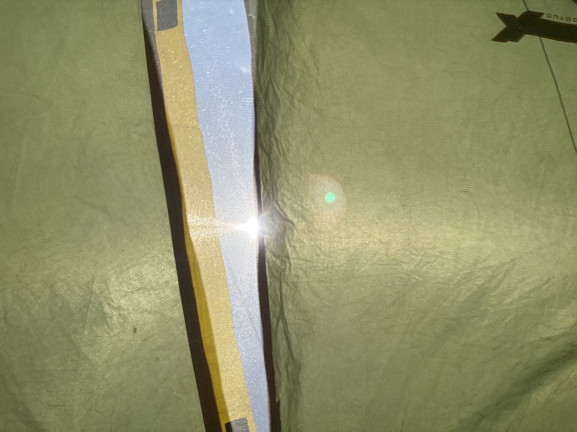 The image shows a close-up interior view of a tent, likely made of olive-green fabric. A strip of light is visible between the tent flaps, revealing a yellow and blue section. Bright light is shining through the gap, creating a lens flare effect. A dark-colored logo is partially visible in the upper right corner. The fabric of the tent is textured with horizontal lines. The location, Khvalynsk, Russia, is not discernible from the visual information provided.