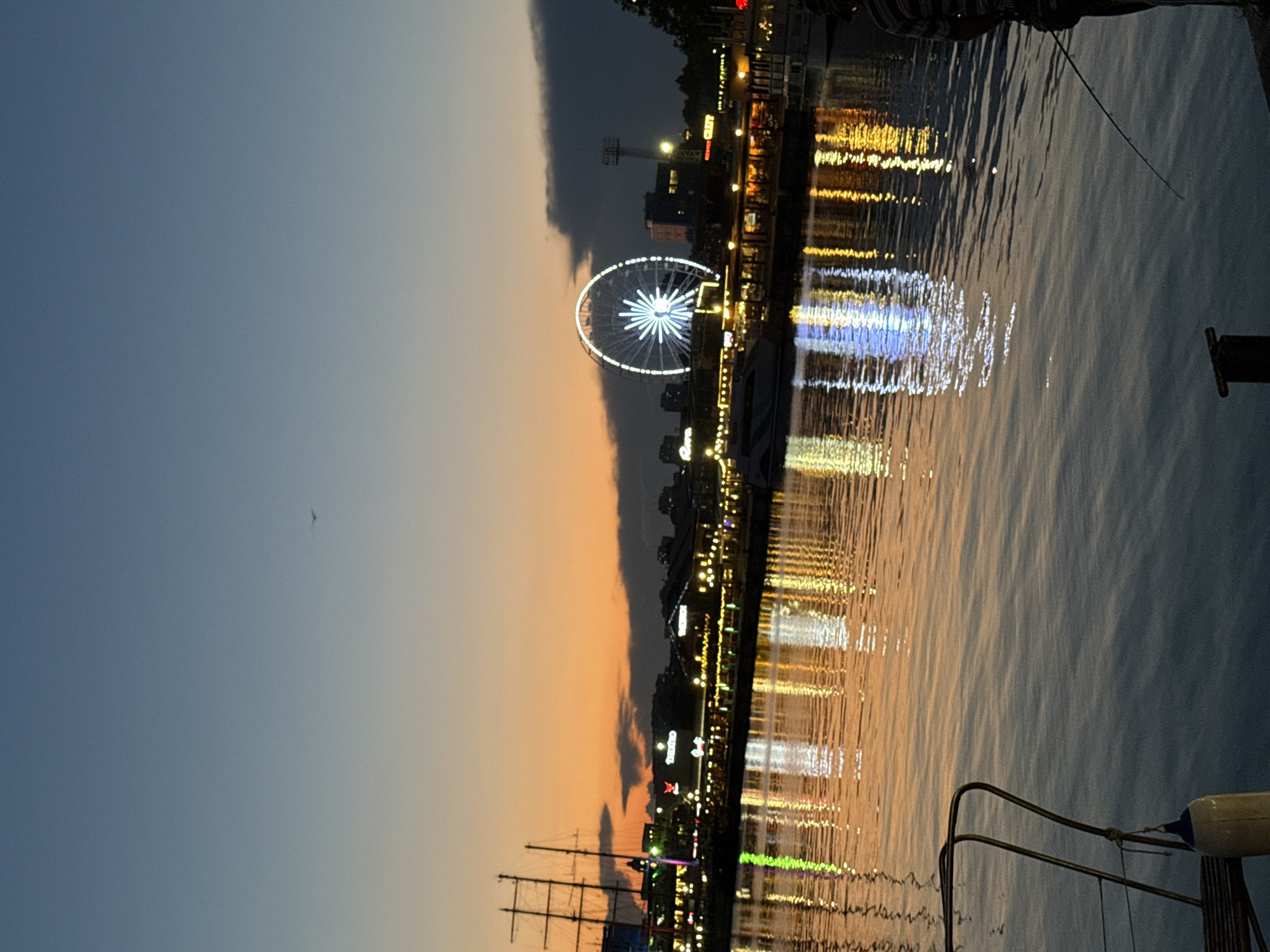 This is a medium shot, taken from a boat, of a waterfront at dusk. The sky is a gradient of deep blue at the top to orange and pink near the horizon. A lone bird is visible in the upper portion of the sky. The water in the foreground reflects the lights of the city, creating a shimmering effect.
In the background, a Ferris wheel is illuminated with white and blue lights. To the left of the Ferris wheel, a tall ship with multiple masts is docked. Several buildings with illuminated windows line the waterfront.
In the lower right portion of the image, a person is fishing from the edge of the boat, with their fishing rod cast into the water. The overall atmosphere is peaceful and serene, with the lights of the city reflecting on the calm water.