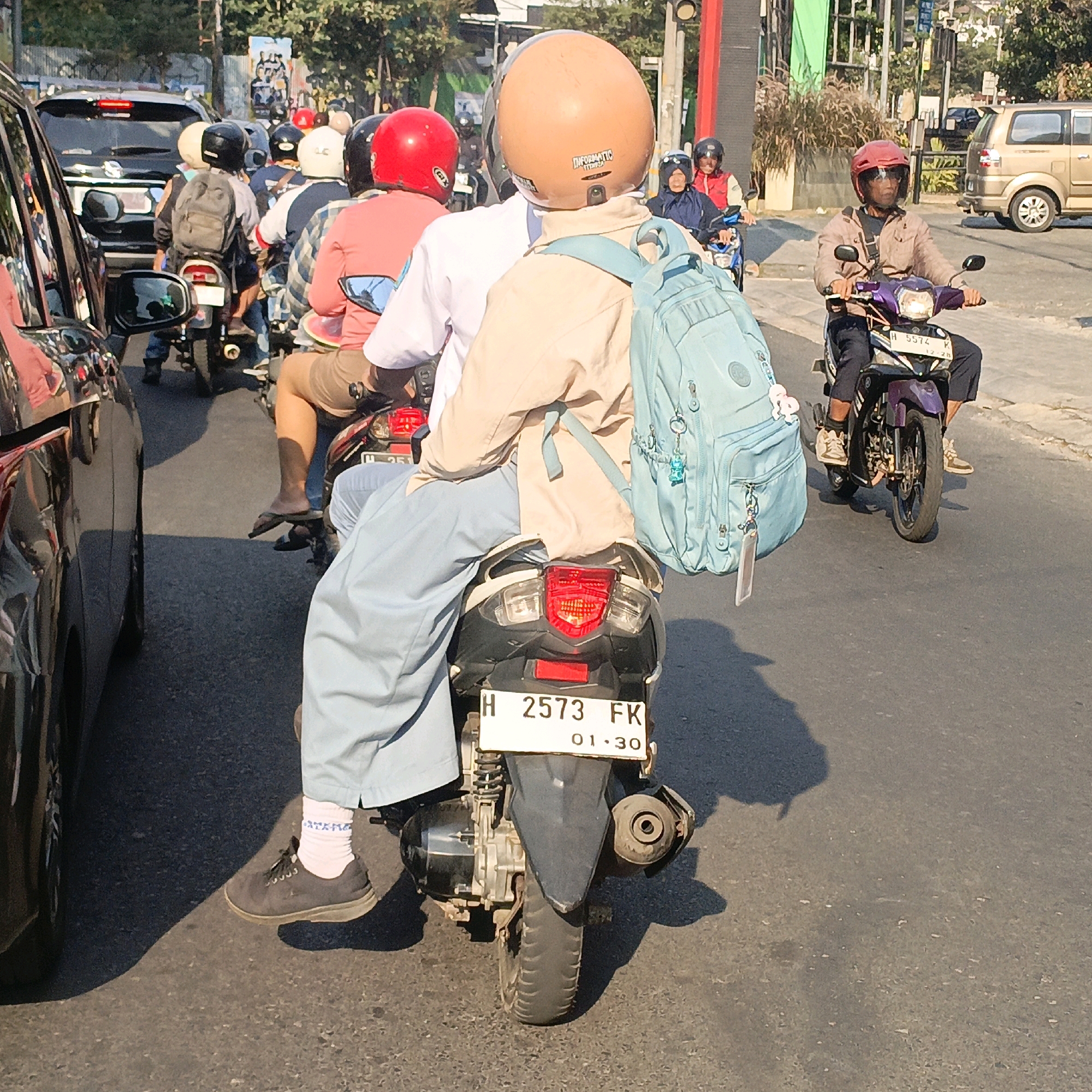 In the city of Salatiga, Indonesia, this image captures a busy street scene with several motorcycles and cars. The primary focus is on a motorcycle in the foreground, carrying two people. The rider, wearing a beige jacket and a light brown helmet, has a light blue backpack strapped to their shoulders. The passenger behind them is wearing a light blue uniform and a beige jacket, also carrying a light blue backpack. Their motorcycle's license plate reads "H 2573 FK" with an expiration date of "01.30".
To the left of the foreground motorcycle, a black car is partially visible, indicating traffic congestion. Further ahead, other motorcycles are seen, with riders and passengers wearing various helmets, including red and black ones. One motorcycle in the middle ground has a rider in a red helmet and a passenger in a pink shirt and brown shorts. Another motorcycle further back shows a rider in a red helmet.
The environment suggests a warm, sunny day, with clear blue skies overhead and shadows cast on the asphalt road. Trees and buildings can be seen in the background, typical of an urban landscape in Indonesia. A gold-colored car is also present on the right side of the image, suggesting a mix of vehicles on the road. The overall impression is one of a typical day on Indonesian streets, with numerous motorcycles navigating the traffic.