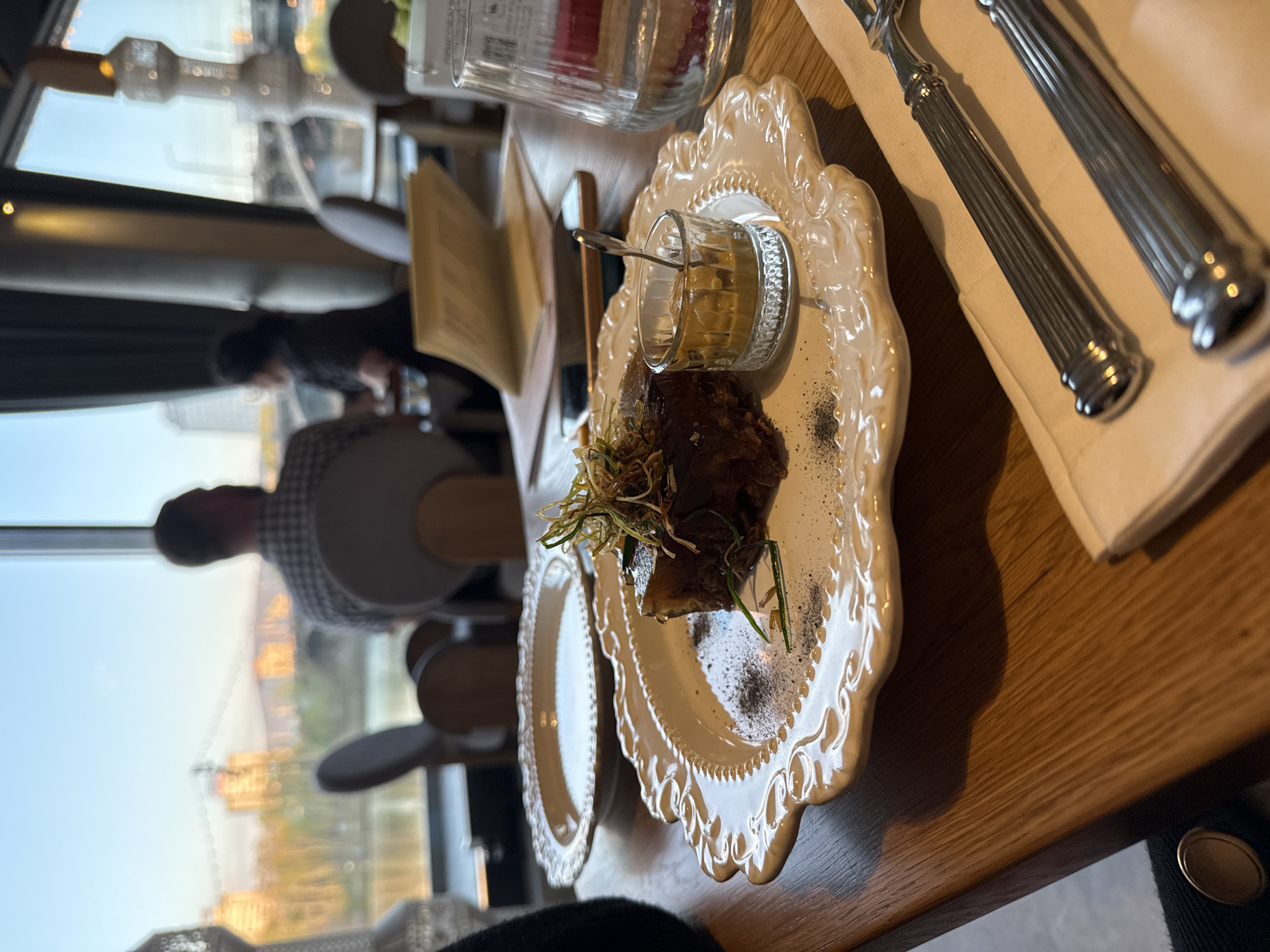 The image captures a dining scene at a restaurant. A wooden table hosts a decorative plate with a dark-colored food item, garnished with herbs and sprinkled with a black powder, possibly pepper. Next to it is a small glass containing a light yellow sauce and a spoon. Cutlery is arranged on a napkin on the table. In the background, blurred figures of diners can be seen near a window that provides a view of a cityscape, possibly Krasnoyarsk, Russia, given the provided context. Buildings and a river are visible through the window. The scene suggests a restaurant setting with a focus on a meal.