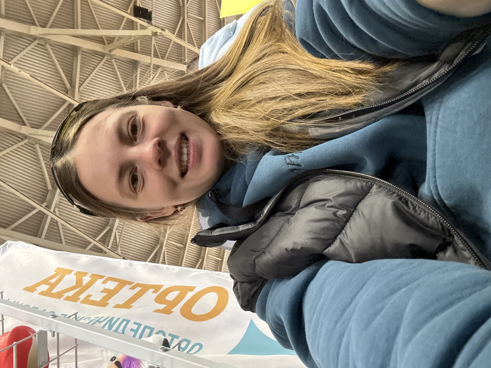 A young woman smiles warmly for a selfie, positioned close to the camera. She has long, light brown hair, a bright expression, and is wearing a blue hooded sweatshirt underneath a dark, possibly black or dark gray, puffer vest. A dark headband or pair of sunglasses rests on top of her head.
The setting is indoors, likely a large public space such as an exhibition hall or arena in Песчаные Ковали, Russia. The background is dominated by a high, light-colored ceiling with a complex geometric pattern of beams and corrugated panels. Below the woman, a white banner is visible, displaying large orange Cyrillic text that, when oriented correctly, reads "ОБЪЕКТ" (meaning "Object" or "Facility"). Below this, smaller text, partially obscured, appears to include "УНИВЕРМАГ" (Department Store). A light blue wave pattern is also visible on the banner. Some items, including a red and white object and a purple and white fluffy item, can be seen on a display rack or shelf just below the banner. The overall impression is that of a person enjoying an event or outing in a spacious venue.