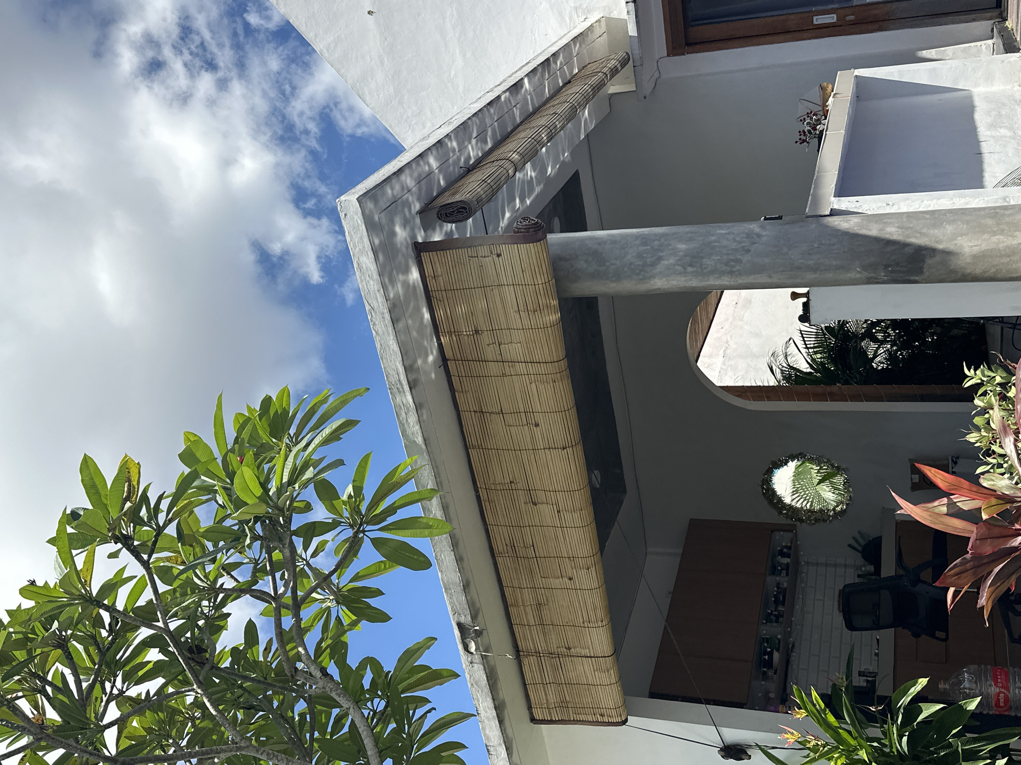 The image shows an upward view of a building exterior, partially obscured by a leafy tree. The sky is visible, with scattered clouds. The building is white with bamboo shades attached. A concrete support beam is in the foreground. The scene contains various plants and building interiors are partially visible, suggesting a domestic setting. The location is Dalung, Indonesia.