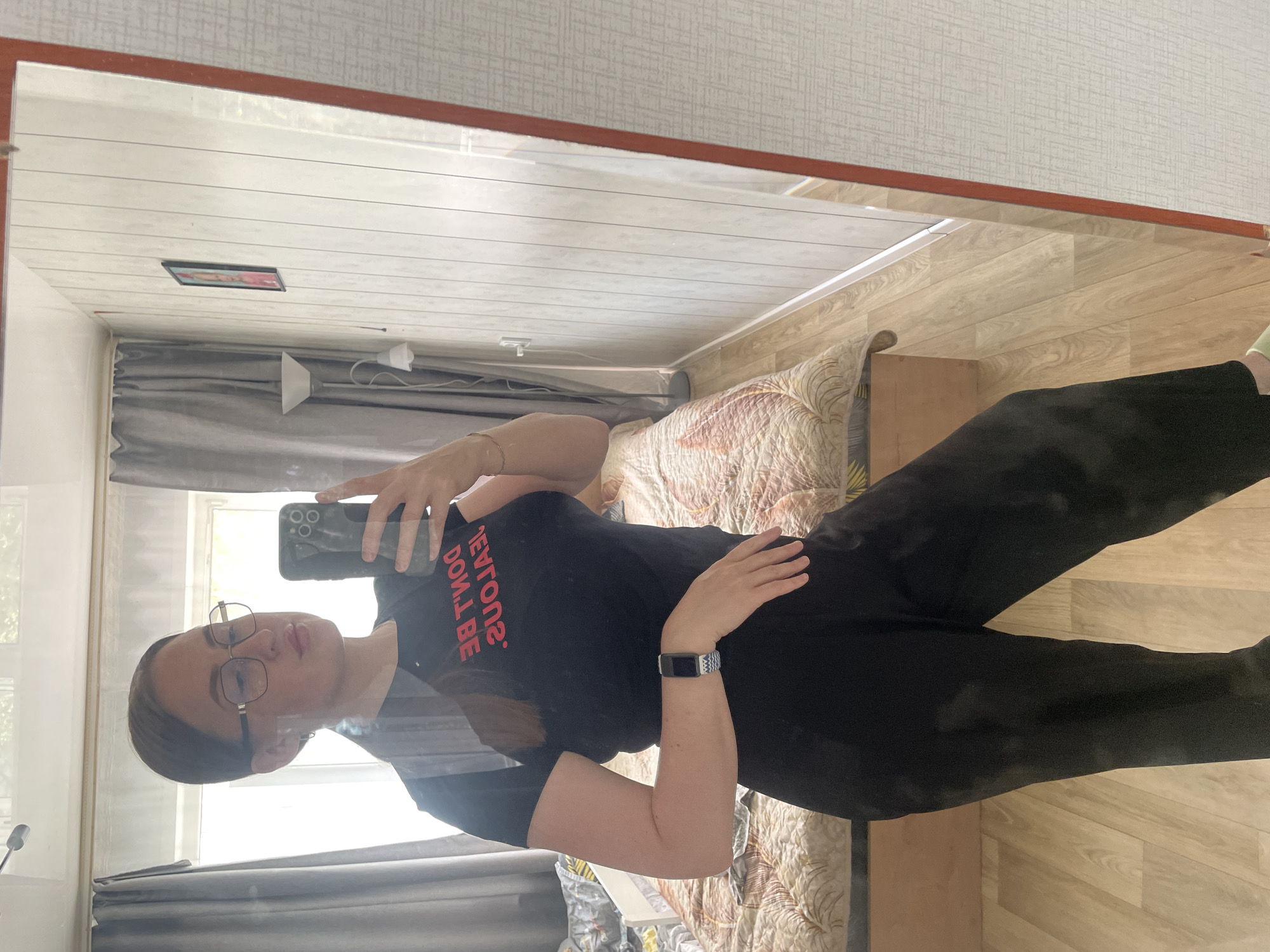 A woman is taking a mirror selfie in a room. She is wearing a black t-shirt with red text that reads "DON'T BE JEALOUS" in reverse, black pants, glasses, and a fitness tracker on her left wrist. The room appears to be a bedroom with a bed with a patterned quilt, curtains, and a floor lamp. A framed picture is visible on the wall in the reflection. The floor is light-colored wood. The overall environment suggests an indoor residential setting, possibly in Сырково, Russia.