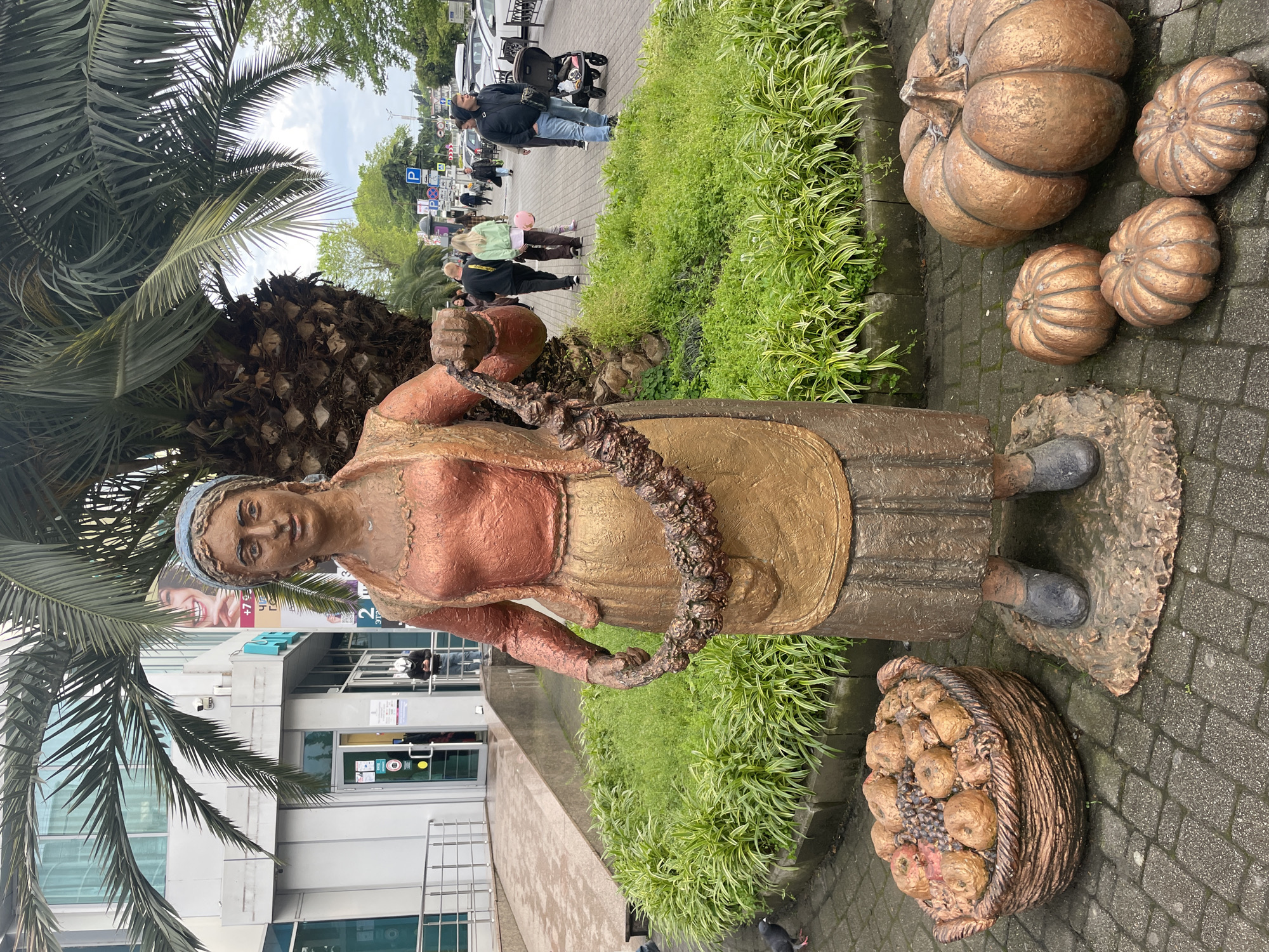 The image depicts a bronze-colored statue of a woman in a traditional dress, possibly a local farmer or vendor, surrounded by decorative pumpkins and a basket of produce. The statue stands on a cobblestone pavement next to a flower bed. A palm tree is visible on the left side of the image. In the background, people are walking on a sidewalk in front of a modern building with glass windows and doors. The location is likely a public space in the city of Sochi, Russia, based on the architecture and vegetation visible in the scene.