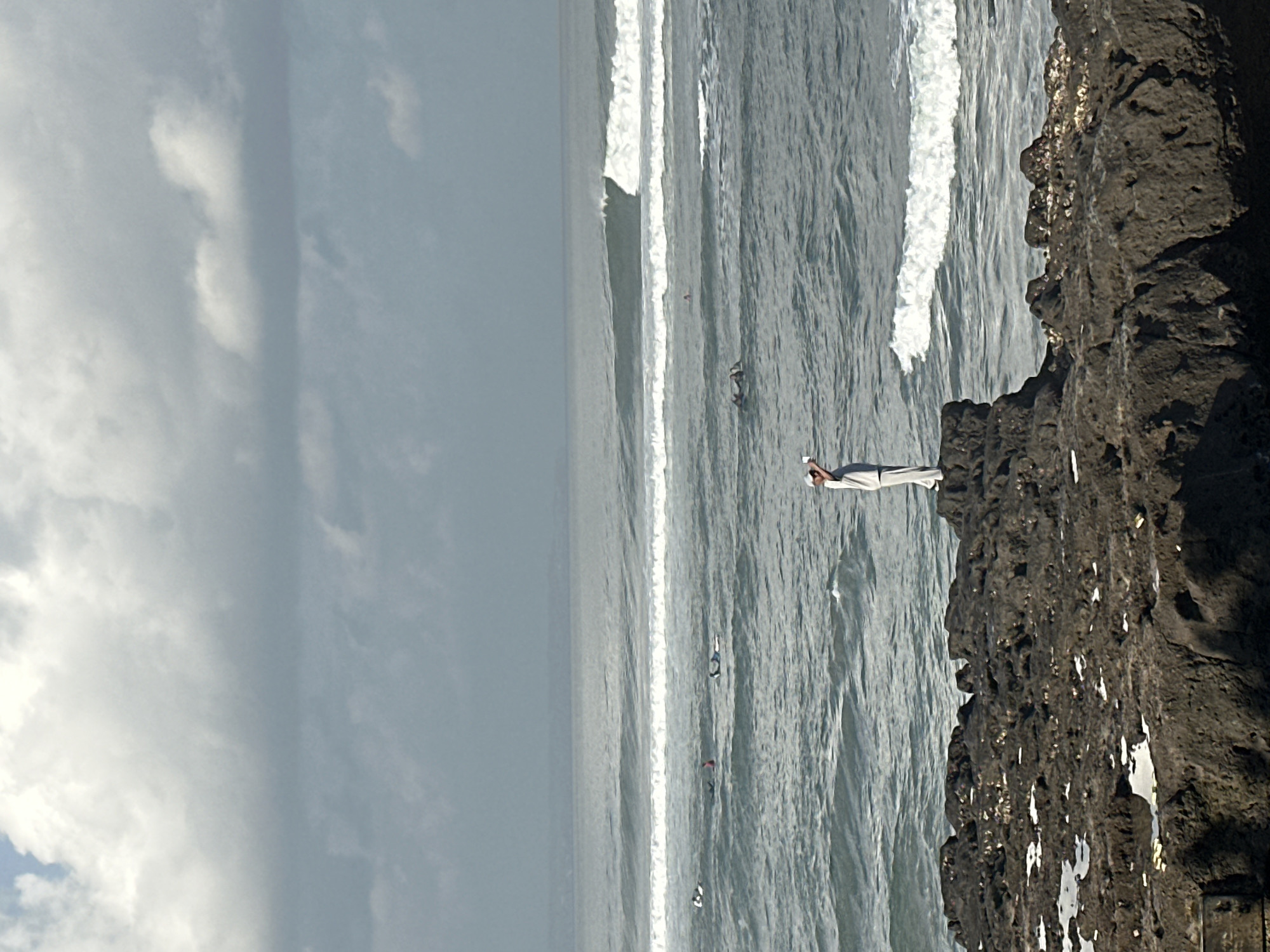 A person in a white outfit stands on a rocky outcrop facing the ocean in North Kuta, Indonesia. The person is looking out at the water and appears to be holding their hand to their head. Several surfers are visible in the ocean, some paddling and others riding waves. The sky is cloudy, and the waves are moderate. The foreground consists of dark, textured rocks with some patches of white.