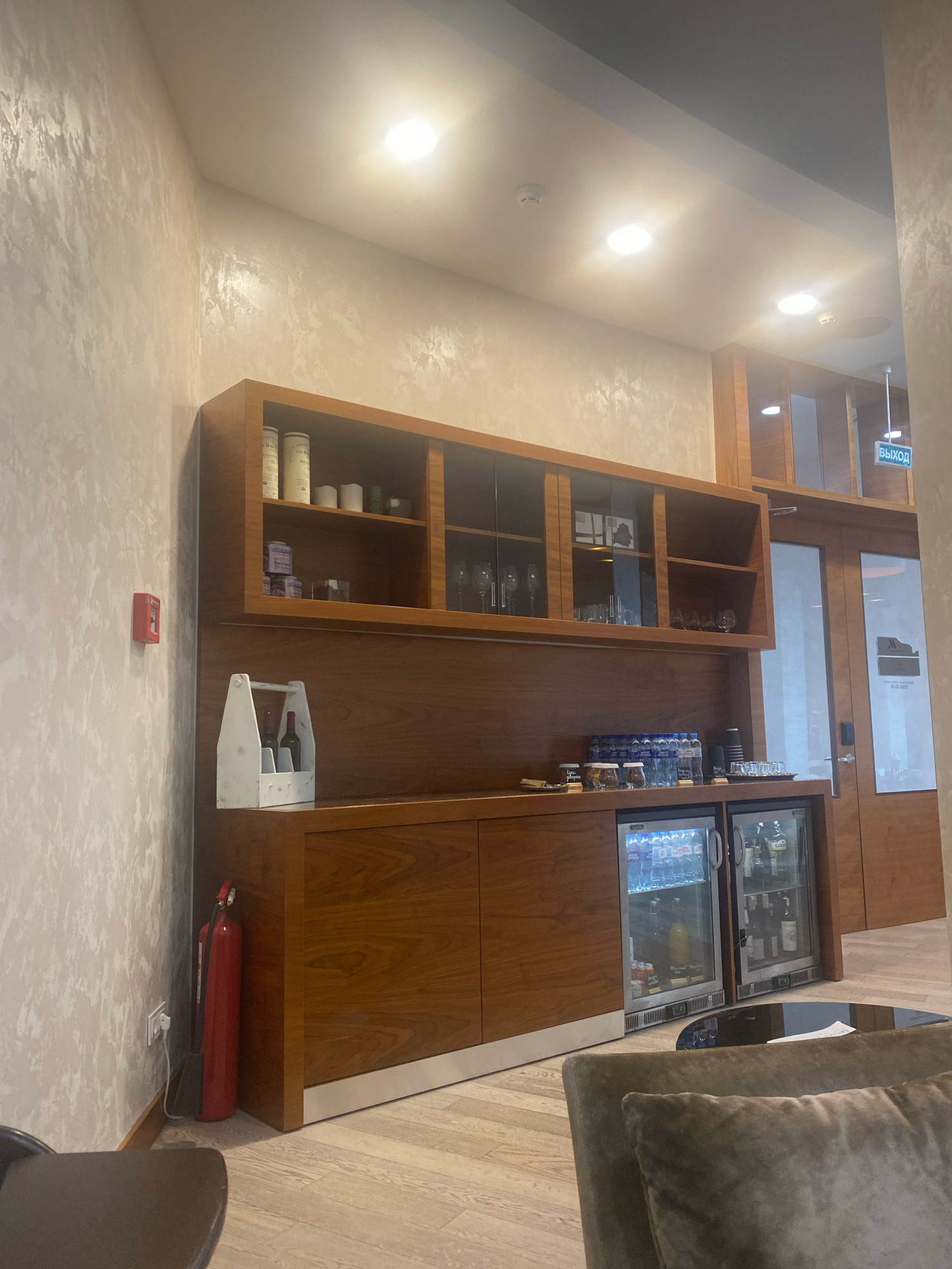 The image shows an interior scene, likely a lounge or waiting area, featuring a wooden cabinet with shelves and two under-counter refrigerators. On the shelves, there are various bottles and items, including what appear to be tea canisters and glassware. The refrigerators contain an assortment of beverages. A white wooden caddy with two wine bottles is placed on the counter. To the left of the cabinet, a red fire extinguisher is visible against a textured wall. Above the cabinets, there is a sign with Russian text that reads "ВЫХОД," meaning "EXIT." The flooring appears to be light-colored wood. The lighting is overhead and bright, with recessed ceiling lights. The overall impression is a clean and functional space. Based on the presence of the "EXIT" sign in Russian and the context of a lounge or waiting area, it is plausible that this scene is located in Minsk, Belarus.