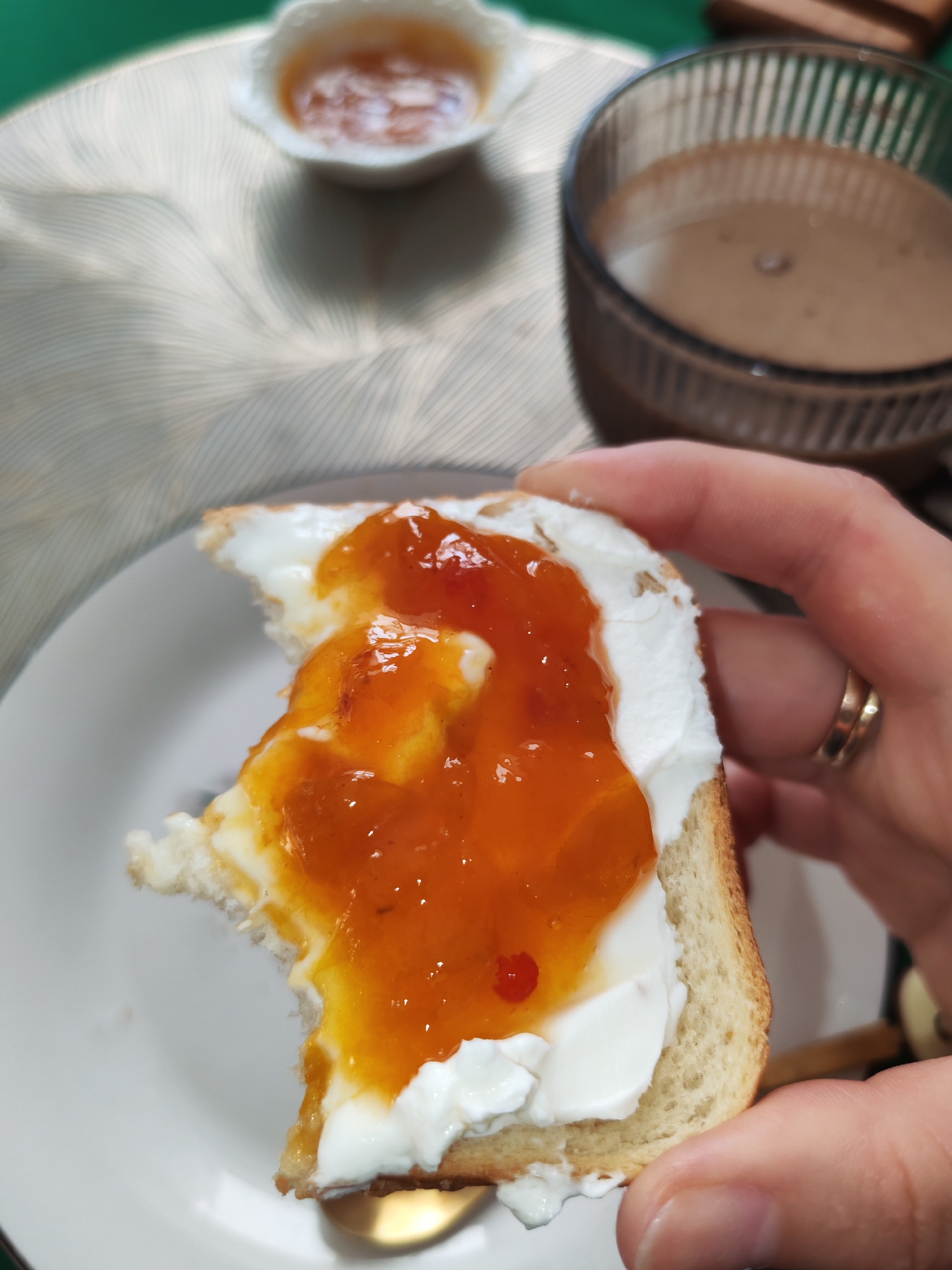 A close-up shot shows a hand holding a slice of bread topped with cream cheese and apricot jam. A small bite has been taken out of the bread. The toast sits on a white plate. In the background, a small white dish contains more jam, and a glass filled with a chocolate-colored beverage sits on a table with a textured tablecloth. The scene appears to be indoors, likely a kitchen or dining area. There is no information available to determine the specific location within Izhevsk, Russia.