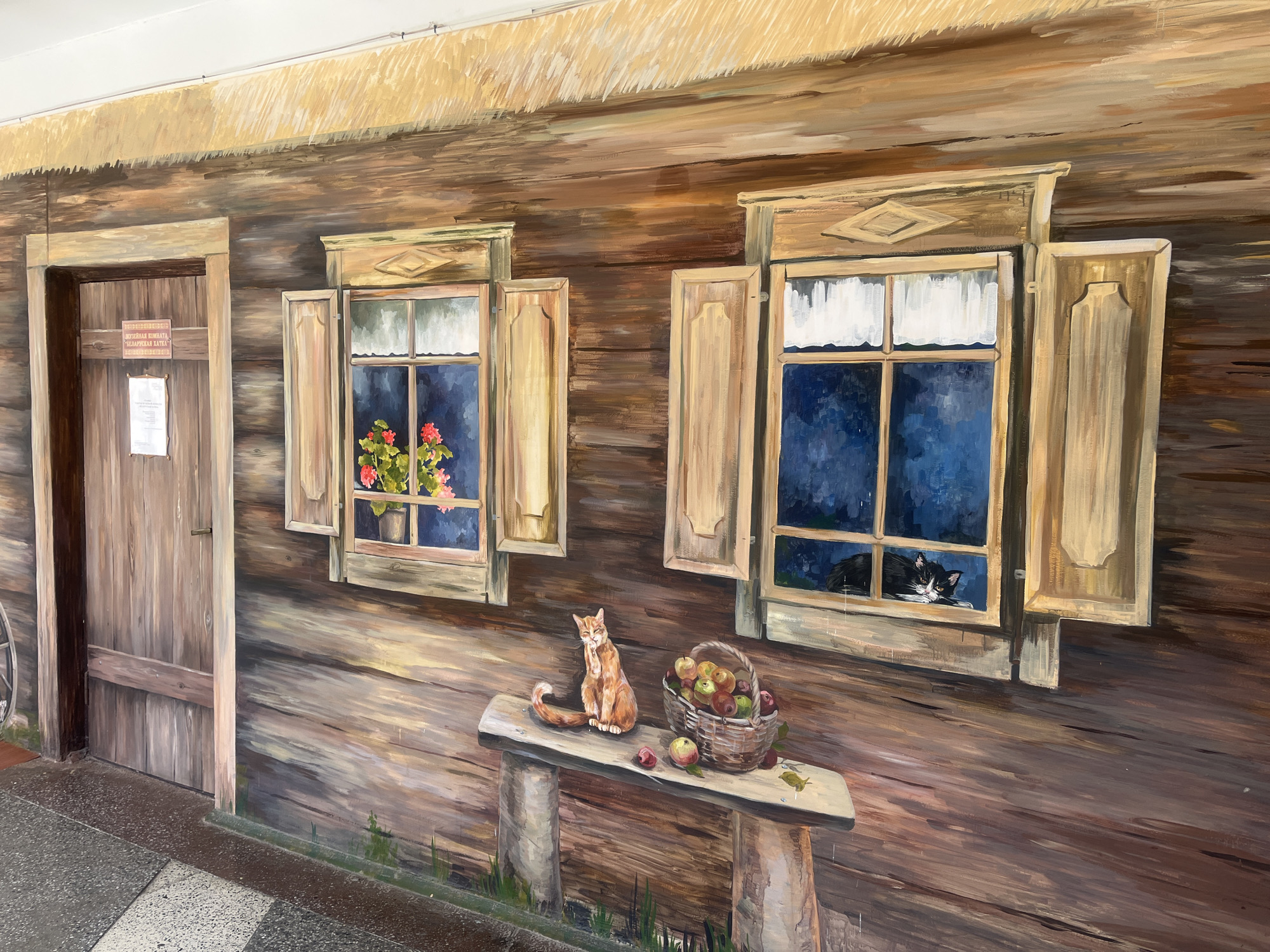 This is a mural painted on the side of a building, depicting a rustic scene. The mural features a wooden facade with two open windows. The left window contains a potted plant with red flowers. The right window shows a black and white cat looking out. In front of the windows is a wooden bench with an orange cat sitting on it, facing left. A basket filled with apples and loose apples are on the bench. The mural is located on the building of the Museum of Belarusian Language in Hrodna, Belarus.