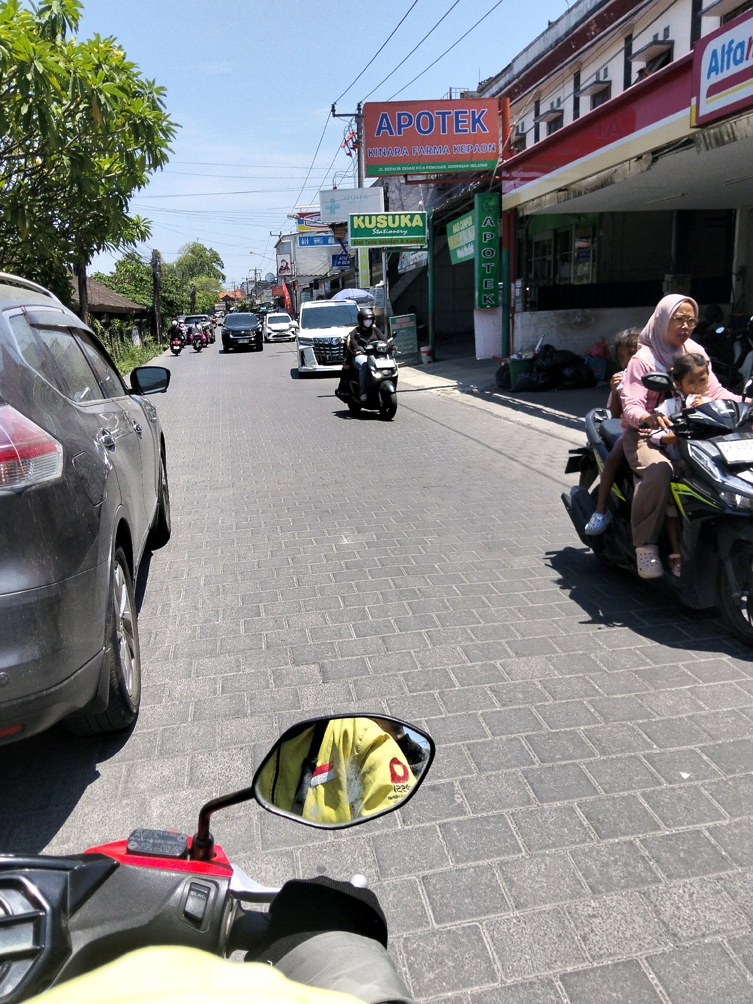 This image captures a street scene in Denpasar, Indonesia, on a sunny day. The perspective is from the viewpoint of someone riding a motorcycle, looking forward down a street paved with gray bricks. To the left, the rear of a dark gray SUV is visible. The street is moderately busy with traffic, including several other motorcycles and cars.
On the right side of the street, there are buildings with shops. A prominent red and white sign reads "APOTEK" (Pharmacy) with additional text below in Indonesian. Next to it is a shop with an "Alfa" sign, likely a convenience store. Further down the street, another green sign reads "KUSUKA Stationery".
Several people are on motorcycles. In the foreground, a woman wearing a pink hijab and a young child are riding on a black motorcycle. Another motorcycle is seen further ahead on the right, with a rider wearing a helmet and a yellow jacket reflected in the rearview mirror of the motorcycle the photo is taken from. The rider of the motorcycle taking the photo is wearing a yellow jacket with what appears to be a small Indonesian flag on the sleeve, also visible in the rearview mirror. The sky is clear and blue.
