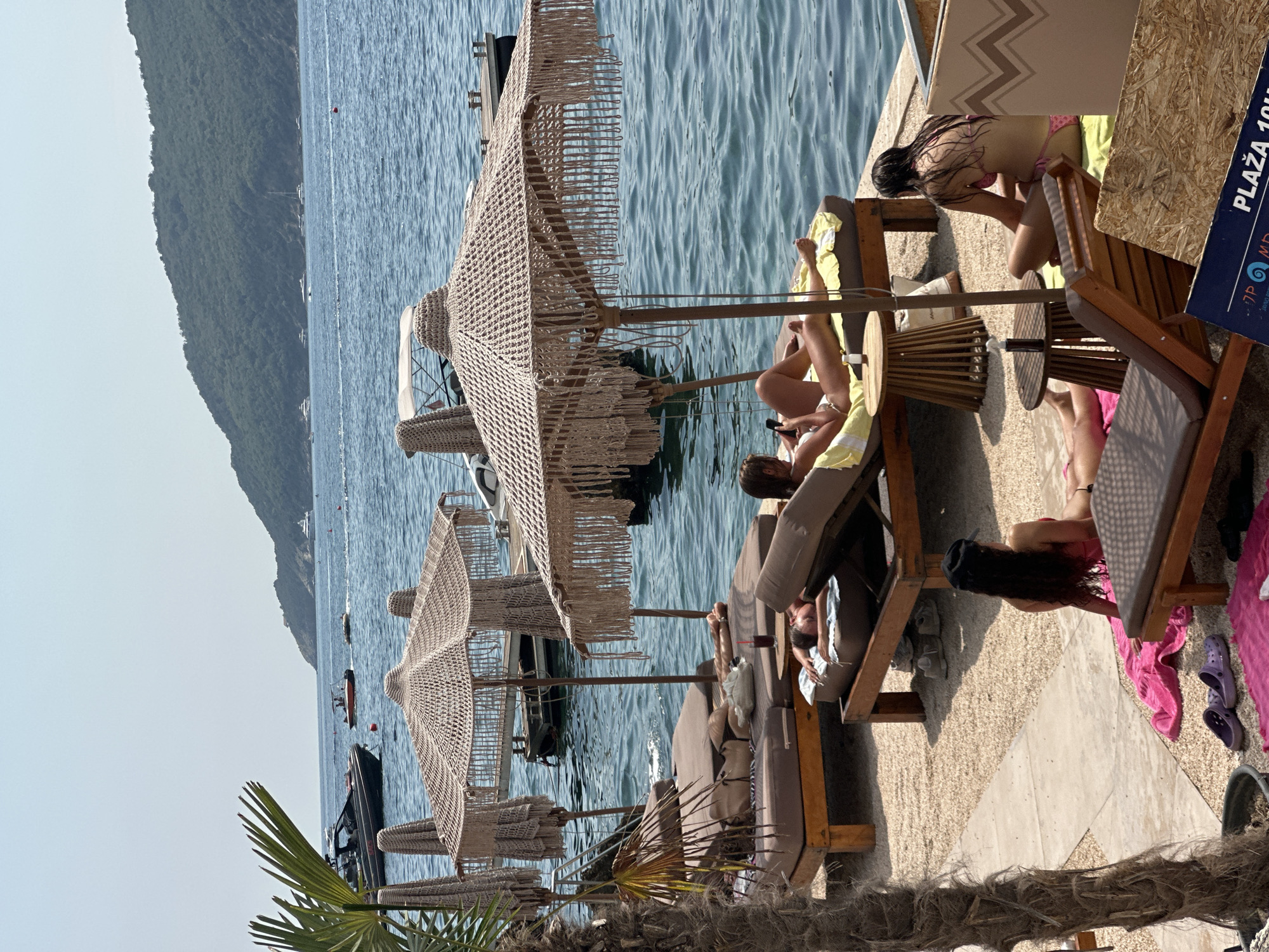 This image captures a lively scene at a beach club in Budva, Montenegro. Several people are relaxing on lounge chairs under thatched umbrellas, enjoying the sunny weather. The beach is covered in light-colored pebbles, and the turquoise water of the Adriatic Sea is visible in the background. Boats and yachts are dotted across the water, and a lush green hill rises in the distance. One person is seen lounging on a chair, scrolling through their phone, while another is reclined with their eyes closed. In the foreground, another individual is seated on a chair, looking away from the camera. The overall atmosphere is one of leisure and relaxation, characteristic of a popular tourist destination.