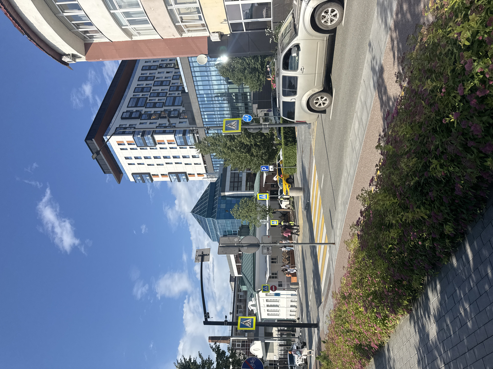 This vertical media file captures an urban street scene in Yekaterinburg, Russia. The image features a mix of modern architecture, including a contemporary glass-fronted building and a multi-story residential complex with numerous windows. A light-colored SUV is parked on the right side of the frame, alongside a pedestrian walkway with a brick-like paving. A lush green bush with small purple flowers lines the walkway, casting dappled shadows on the ground.
Several pedestrian crossings with yellow and white striped markings are visible on the road. Road signs, including a yellow diamond-shaped sign with a pedestrian symbol and a blue circular sign with a bicycle symbol, are mounted on street poles. People can be seen walking on the sidewalk and crossing the street, suggesting a moderate level of activity. The sky is a clear, vibrant blue with a few scattered white clouds, indicating fair weather. The lighting suggests it is daytime, likely late morning or afternoon given the angle of the shadows. The overall impression is one of a well-maintained urban environment on a pleasant day. No discernible text is present in the image.