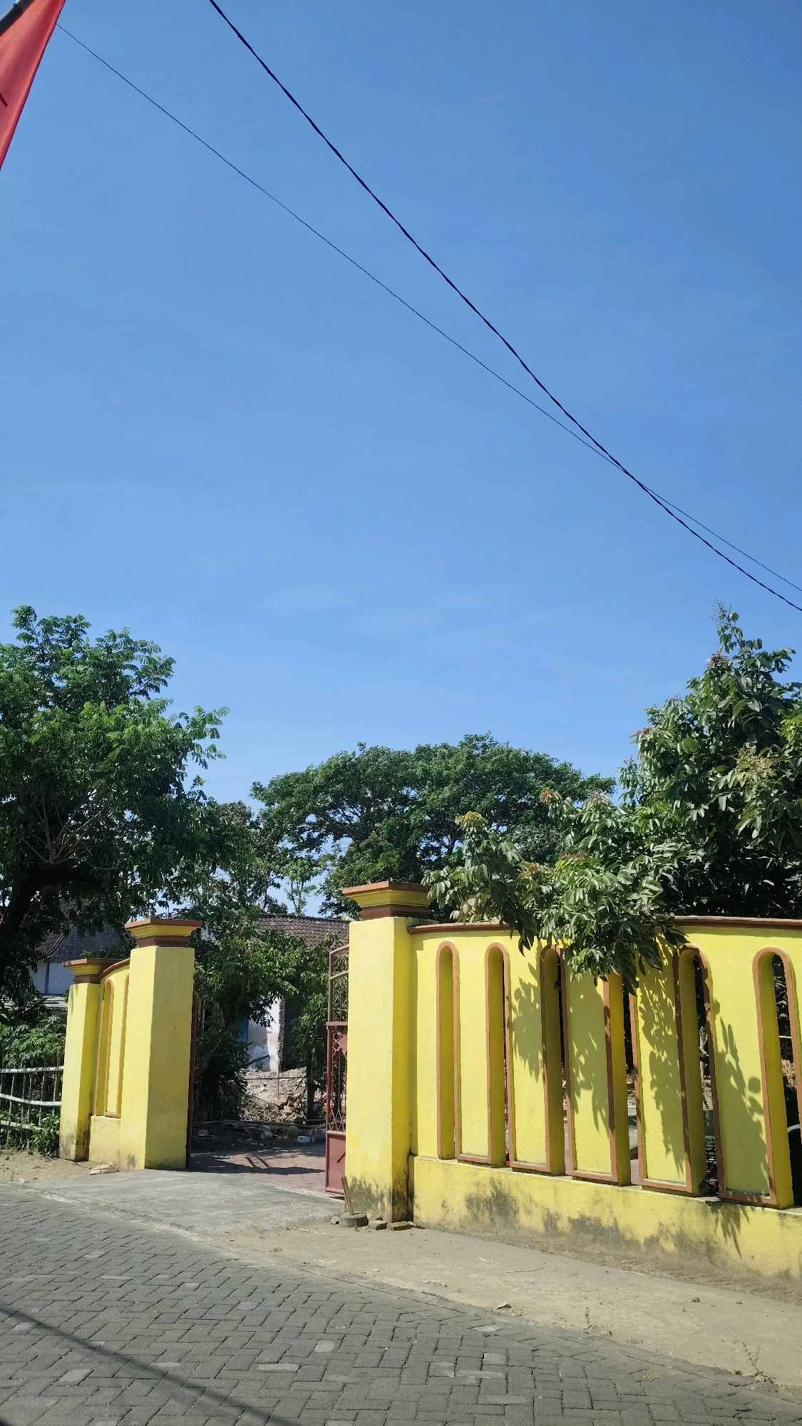 The image captures an exterior view of a property entrance in Jabon, Indonesia. A bright yellow gate, adorned with decorative arched openings, stands prominently. Flanking the gate are two substantial yellow pillars, topped with reddish-brown caps. Beyond the gate, a glimpse of a building with a tiled roof is visible, surrounded by lush green trees. The pathway leading to the gate is paved with grey rectangular bricks, while the area beside the gate appears to be a dirt and sand surface. Overhead, a clear blue sky is bisected by two dark power lines, and a sliver of a red flag or banner is visible in the upper left corner. The scene is illuminated by bright daylight, casting shadows on the yellow wall and the ground.