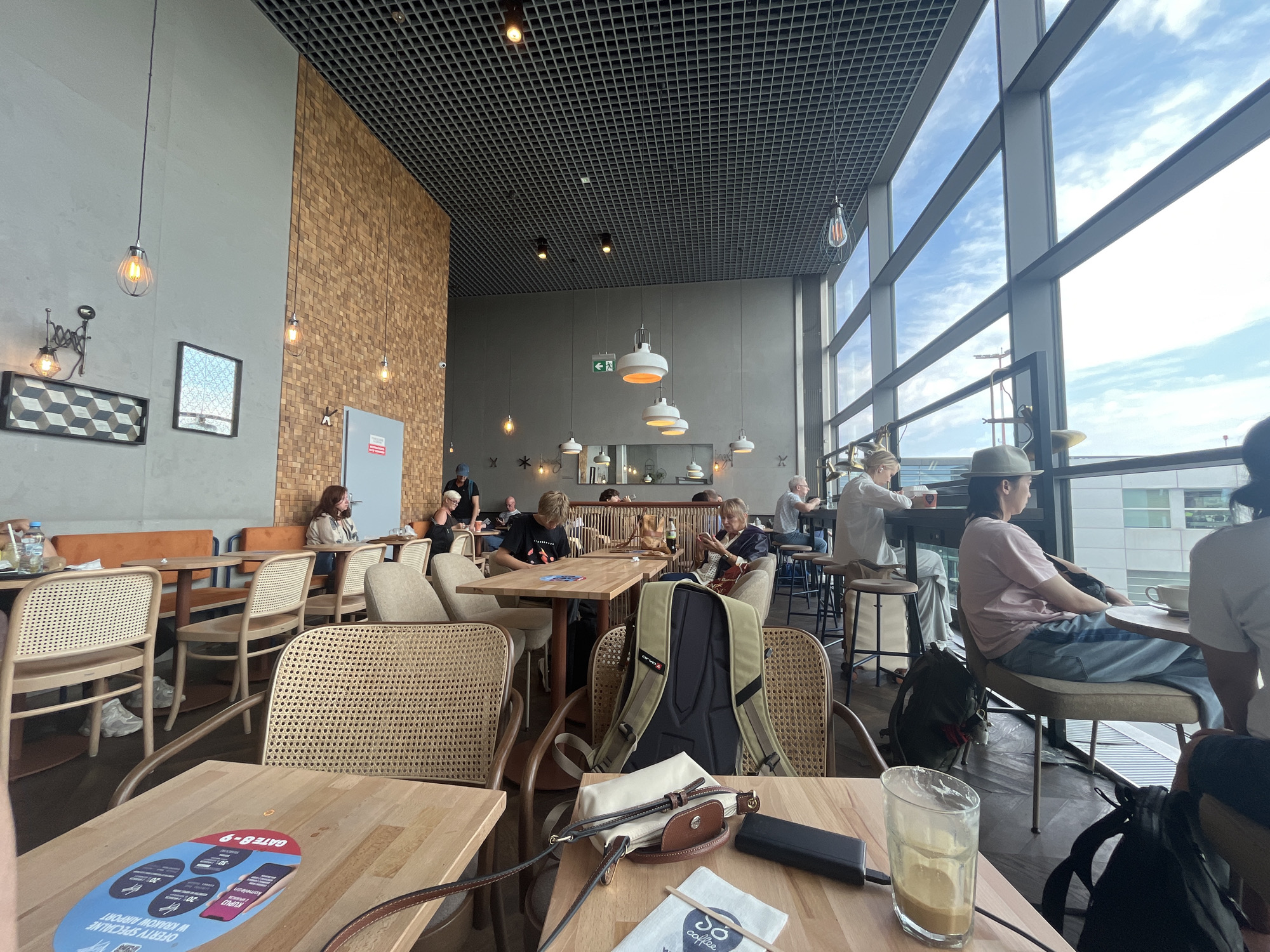This is an indoor photograph of a modern cafe with large windows overlooking an airport terminal in Balice, Poland. The cafe features wooden tables and chairs, some with woven rattan backs, and a large wood-paneled wall. Several people are seated at tables, some dining, some working on laptops, and others conversing. The lighting in the cafe is a mix of natural light from the windows and artificial pendant lights. In the foreground, a wooden table has a beverage glass, a small bag, and a power bank on it, along with a flyer for Krakow Airport. The background reveals more seating areas, other patrons, and a glimpse of the airport's tarmac and aircraft through the expansive windows.