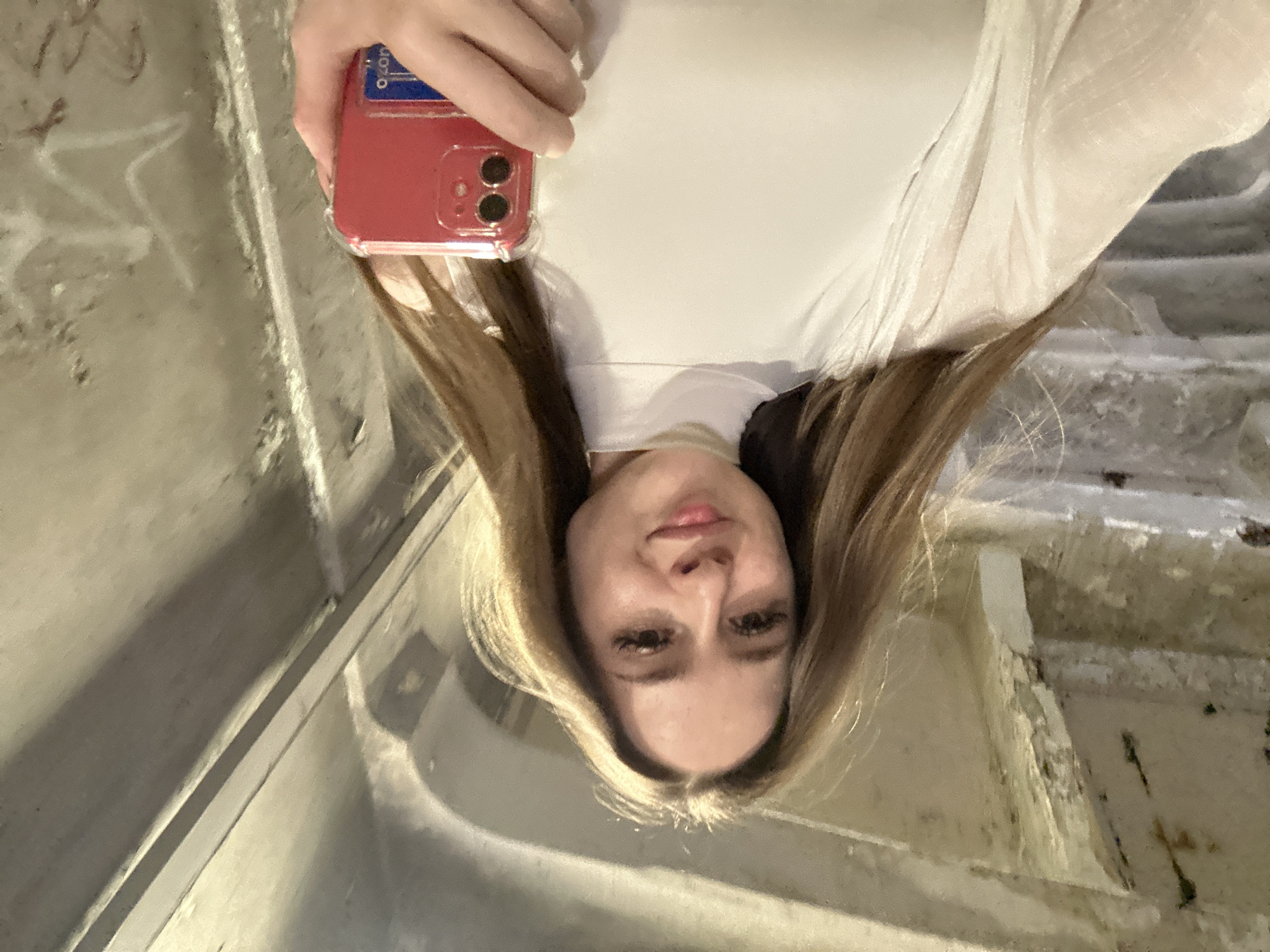 A young woman with long, light brown hair takes a selfie in a dimly lit, industrial-looking space with concrete walls and overhead structures. She is wearing a white, high-necked top and a sheer white outer garment. She holds a red smartphone in her right hand, with a clear protective case. The background appears to be a rough, textured concrete wall with some graffiti visible. The lighting is somewhat harsh, casting shadows on the walls. The perspective is from a low angle, looking up at the woman.