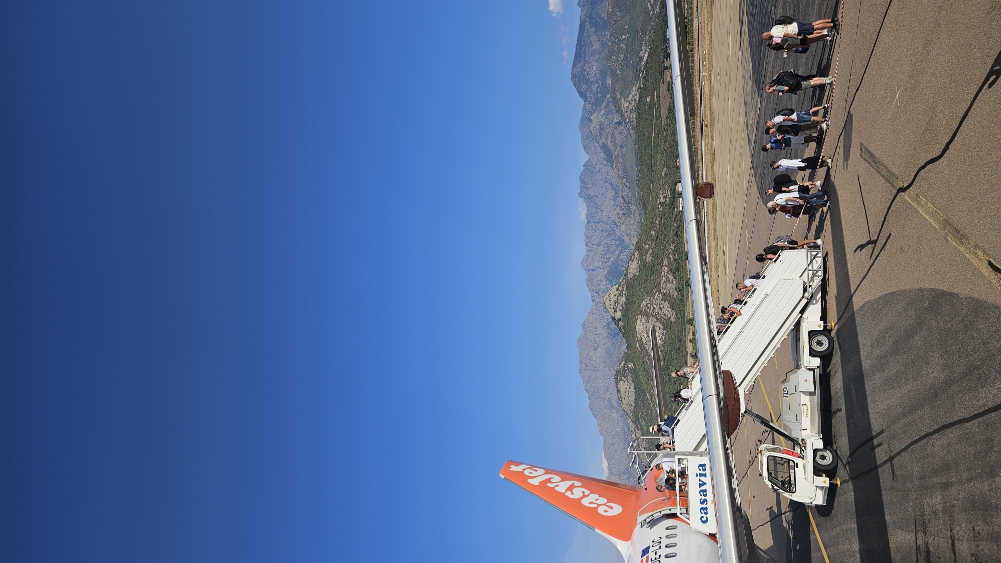 An EasyJet airplane with the registration F-G LOC is parked at an airport with a mobile staircase attached for passengers. People are boarding the aircraft via the stairs. The plane has "casavia" written on its fuselage. In the background, mountains and a clear blue sky are visible. The scene appears to be at Calvi Sainte-Catherine Airport (CLY) in Calvi, France, given the presence of the EasyJet plane and the mountainous landscape characteristic of Corsica.